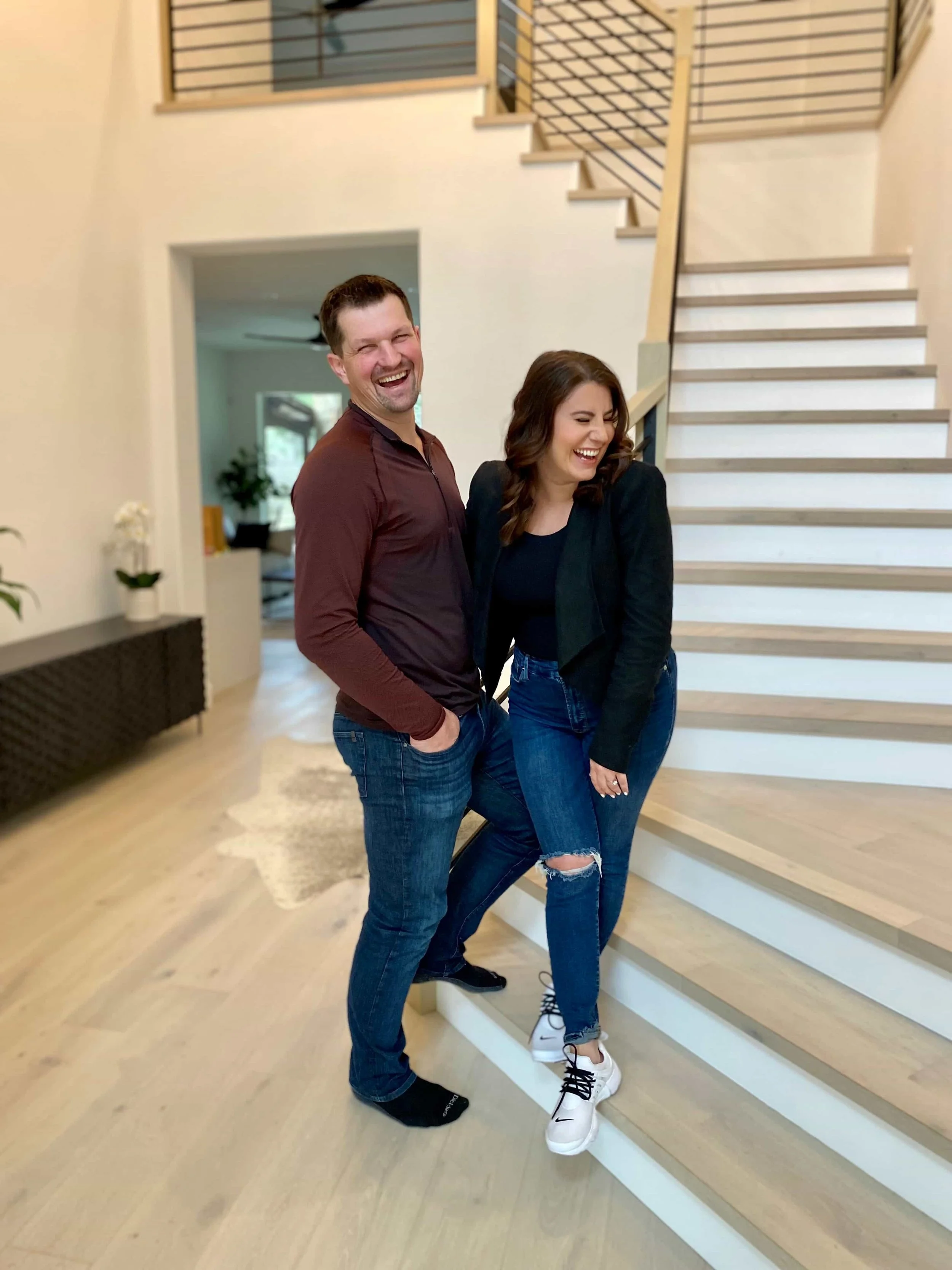 Two happy people, a man and a woman, are standing inside a modern house near a staircase, smiling and laughing.