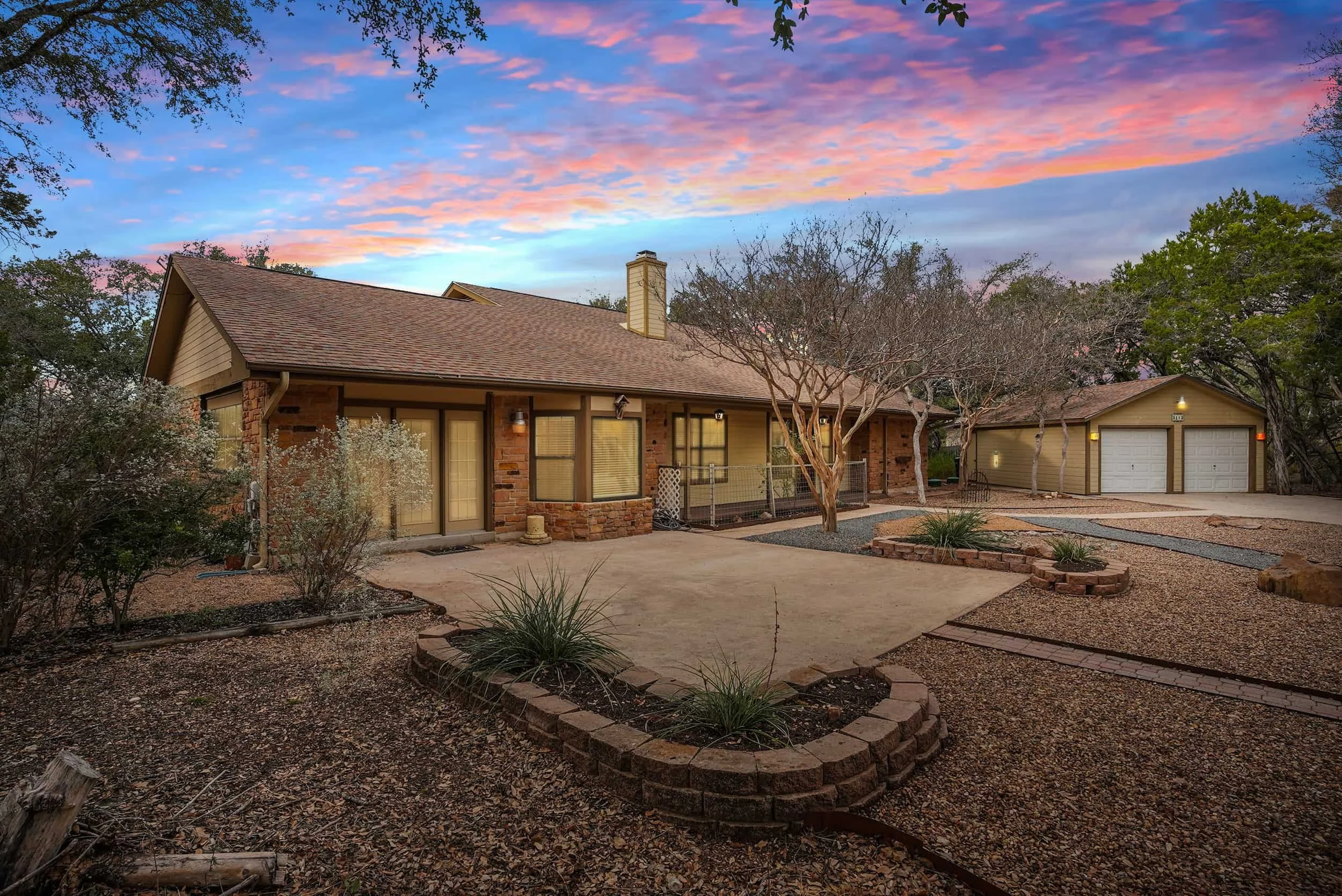 Ranch-style home exterior in Austin, Texas at sunset with warm curb appeal, curved gravel driveway, rock landscaping, and mature oak trees.