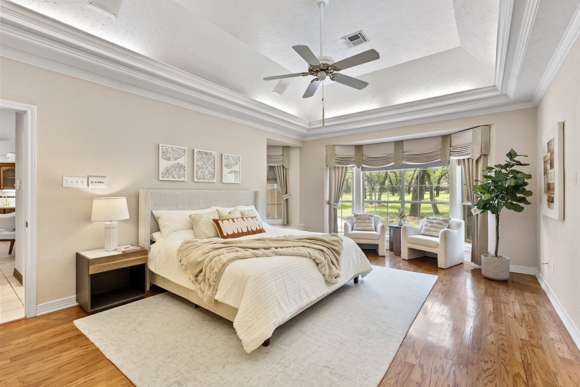 Primary bedroom with a tray ceiling, hardwood floors, and a sitting area overlooking mature oak trees.