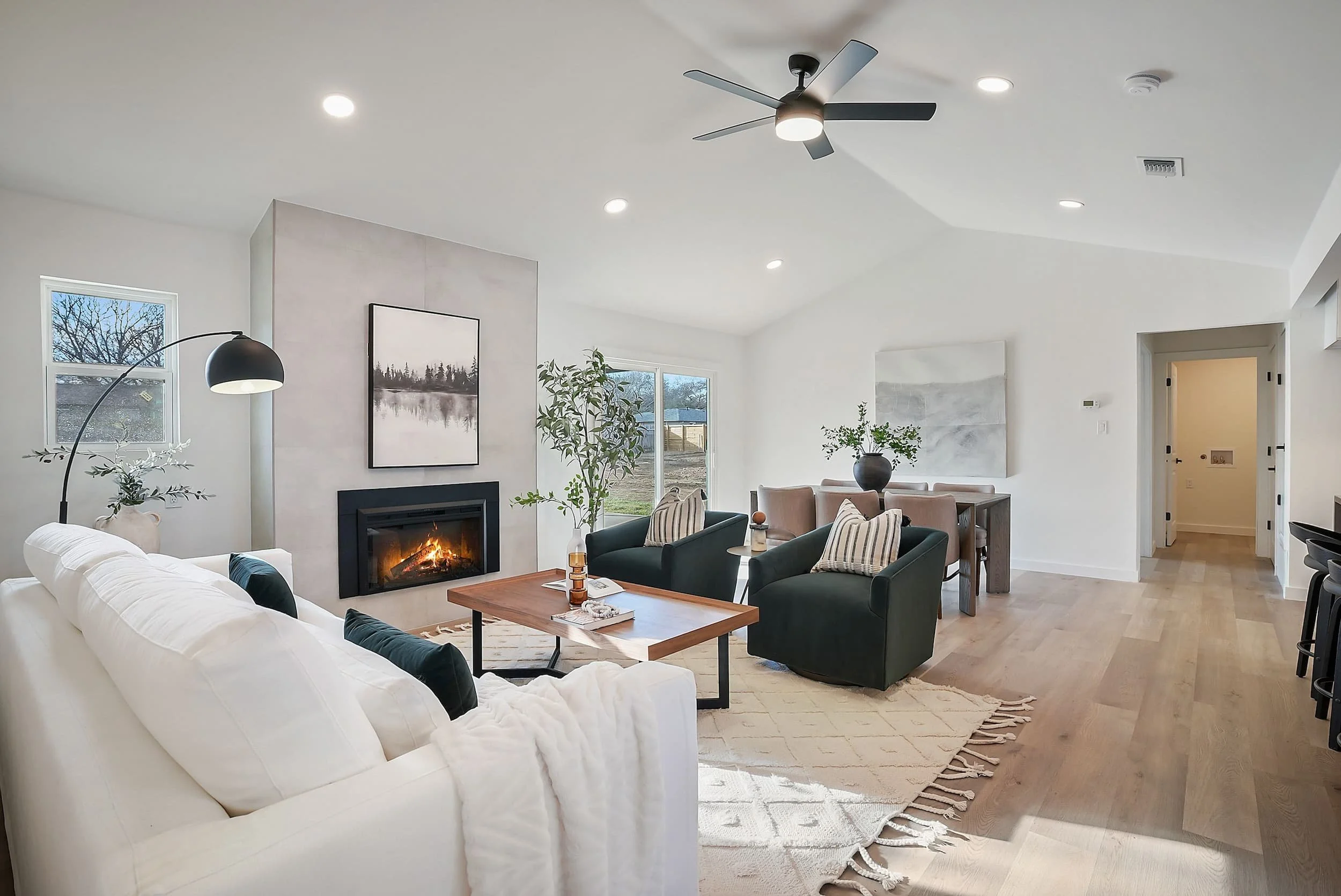 Modern open-concept living room with white sofa, gas fireplace, dining table, ceiling fan, and large windows in neutral tones.