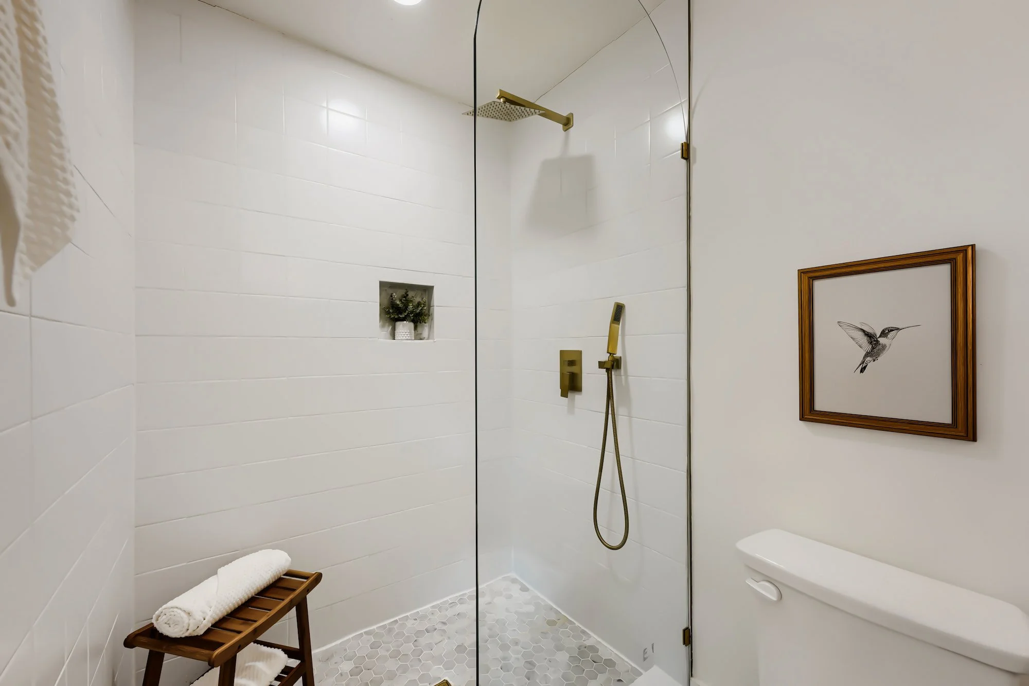 Staged primary bathroom with a glass walk-in shower, white subway tile, brass fixtures, and a wood bench holding rolled towels.