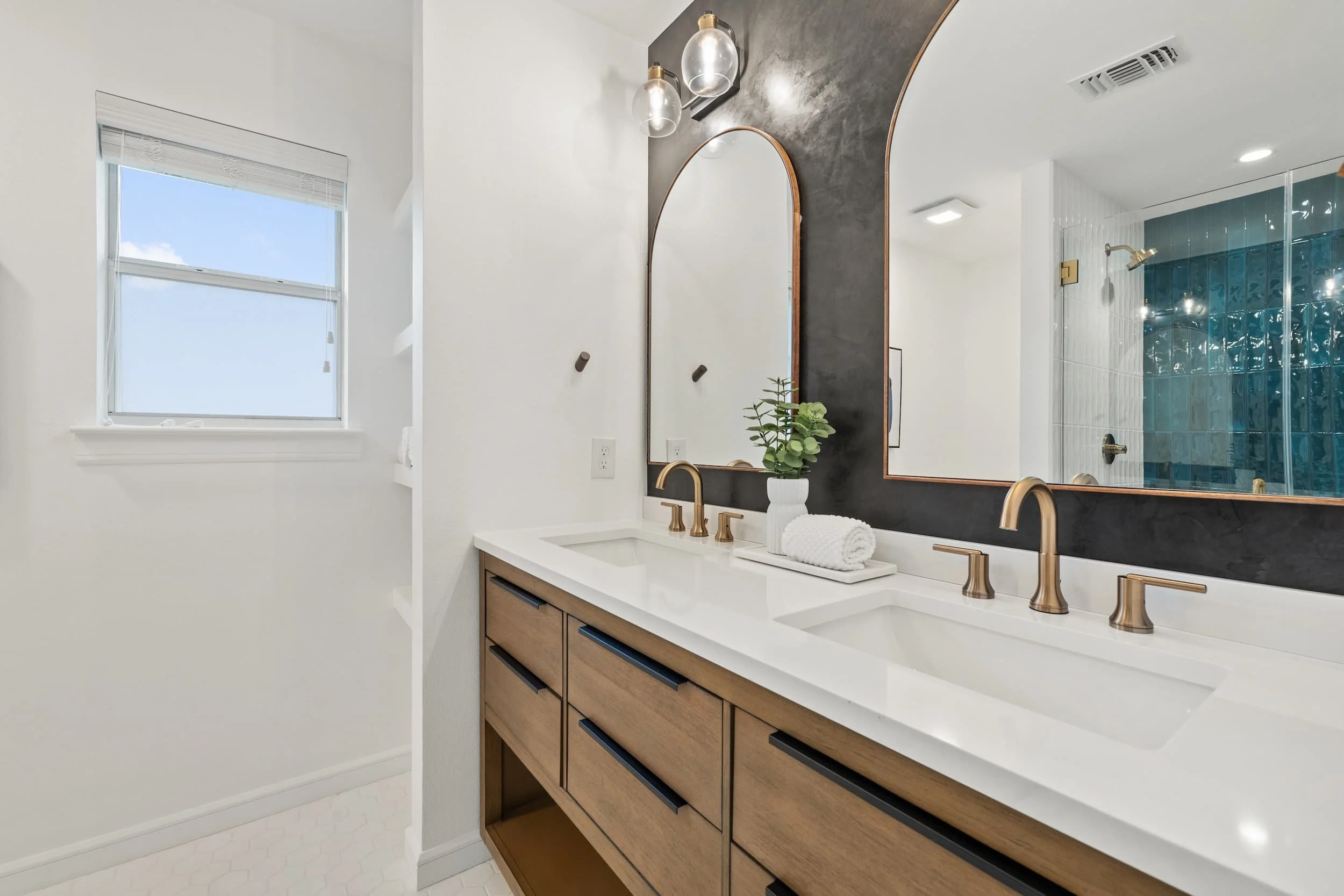 Stylish bathroom with a double vanity, wood cabinetry, gold faucets, large mirrors, and a glass-enclosed shower with blue tile accents.