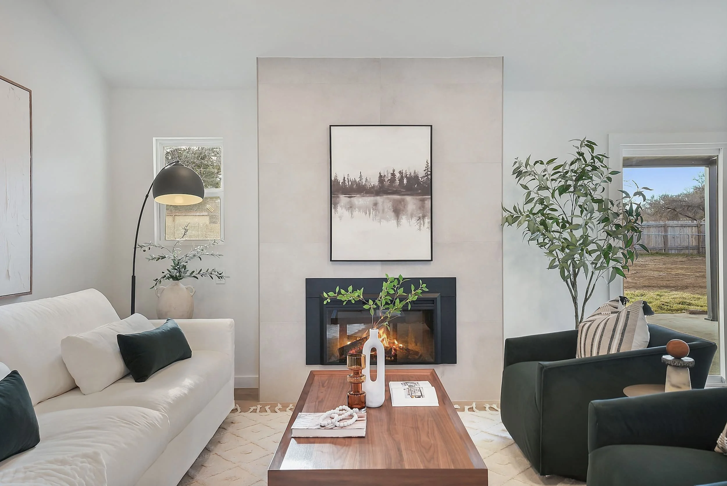 Minimalist living room with black-framed fireplace, tall stone surround, landscape artwork, cream sofa, green chairs, walnut coffee table, and large windows.