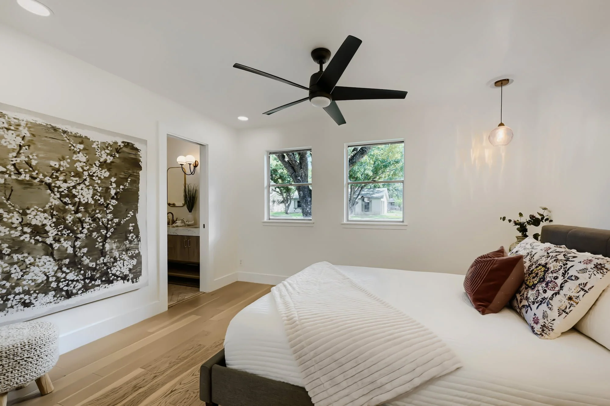 Staged primary bedroom with an upholstered bed, large blossom canvas, and twin windows, opening to an ensuite bathroom.