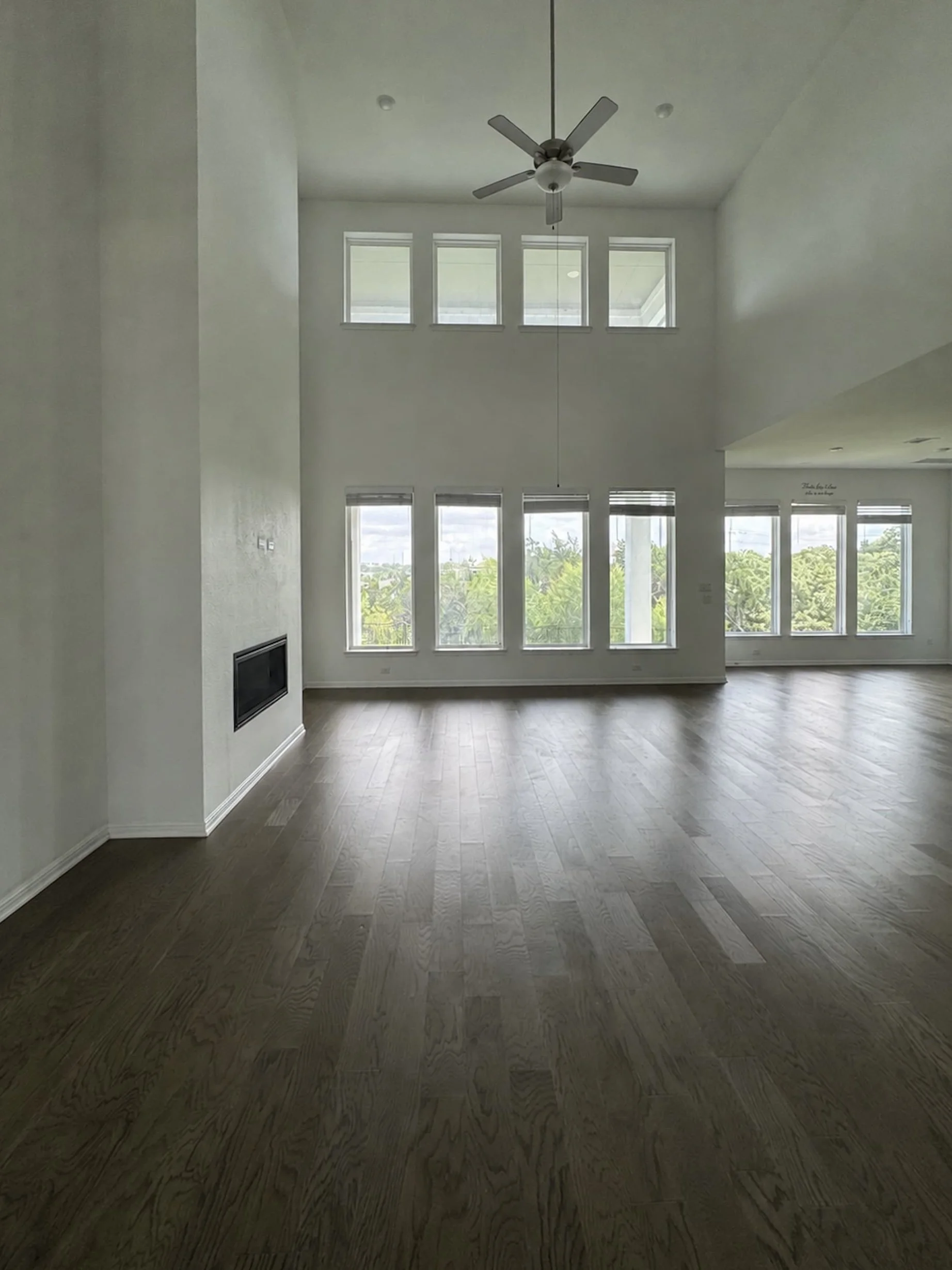 Empty two-story great room with a vaulted ceiling, ceiling fan, two rows of tall windows bringing in natural light, dark hardwood floors, and a linear fireplace on the side wall.