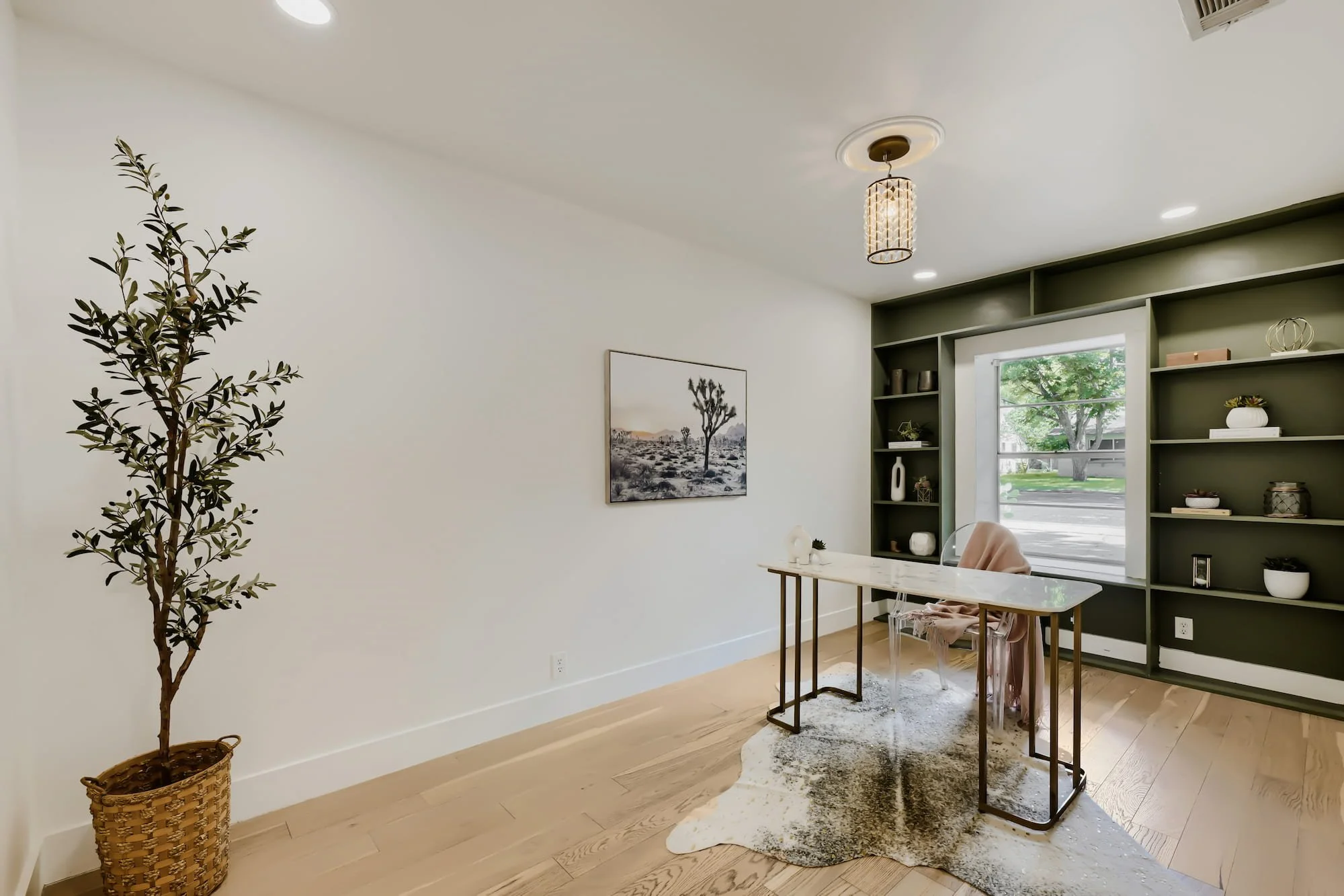 Staged home office with a marble desk, acrylic chair, and olive green built-in shelves framing a window, on a cowhide rug.