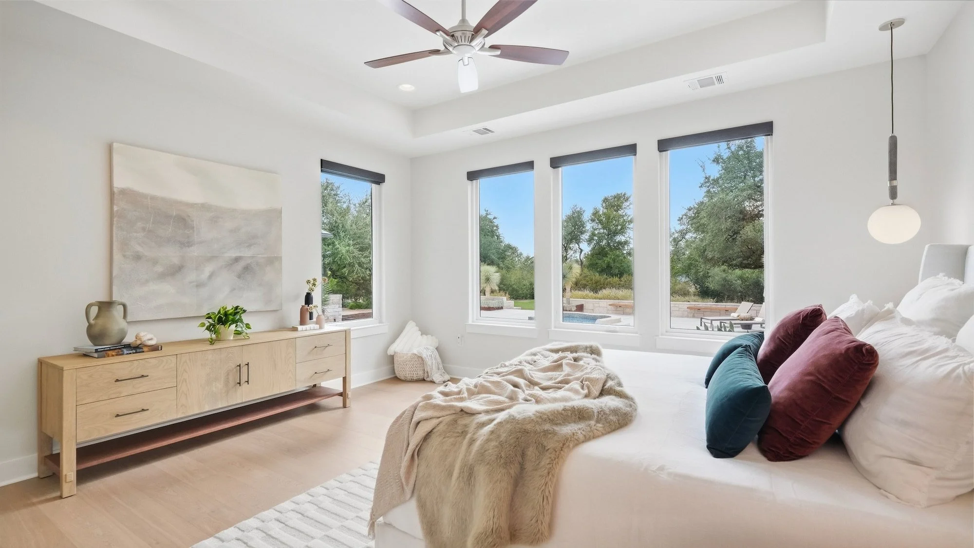 Primary bedroom staged with velvet pillows and an oak dresser, with tall windows overlooking the pool.