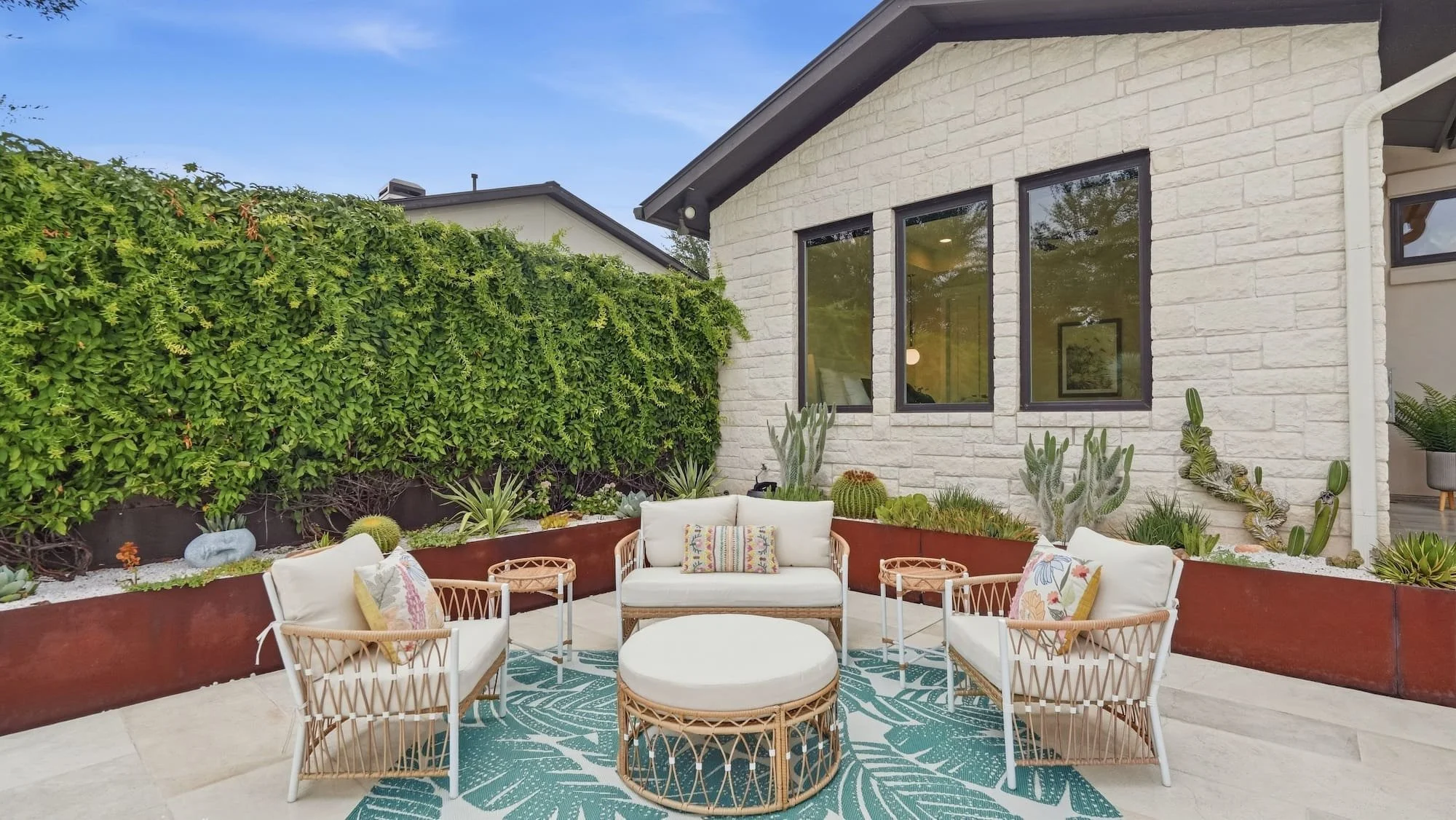 Patio staged with rattan seating and a tropical leaf rug, framed by cactus planters and a vine-covered wall.
