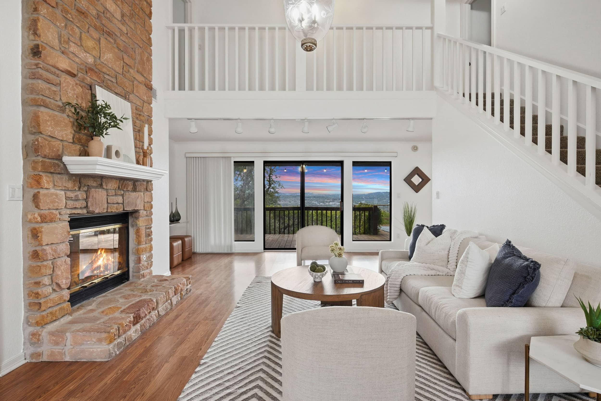 Professionally staged living room with a tall stone fireplace, white sofa, and vaulted ceiling overlooking a deck with a sunset view.