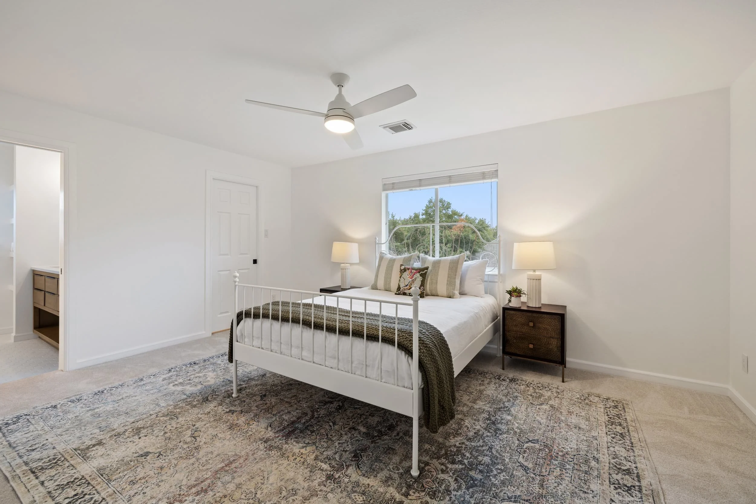 Spacious bedroom with a white metal bed, matching nightstands and lamps, a large window, and soft neutral tones, with an en suite bathroom visible.