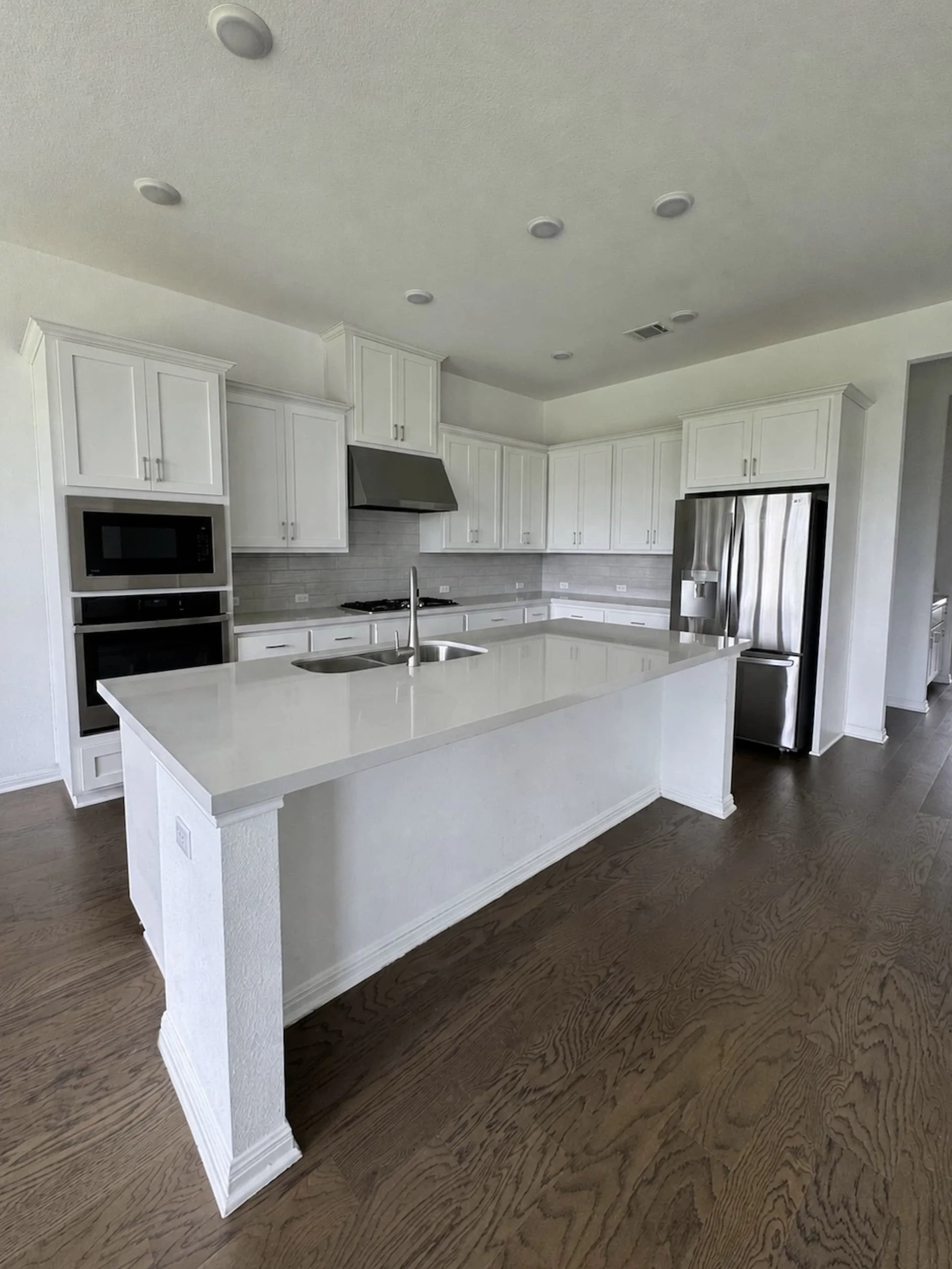 Vacant kitchen with white shaker cabinetry, a gray subway tile backsplash, stainless steel appliances including a built-in microwave and double oven, and a large quartz-topped center island with a sink.