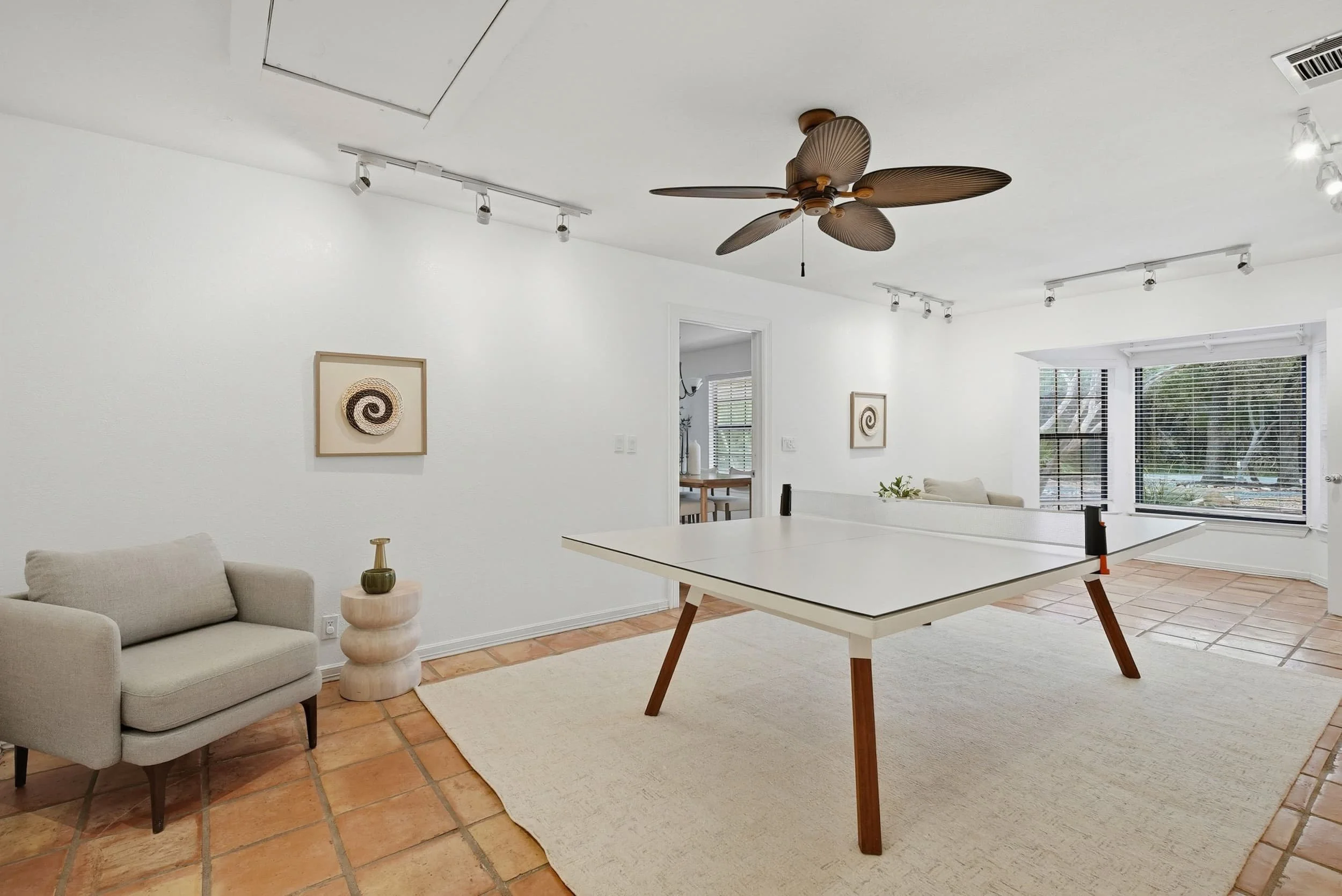 Modern rec room featuring a white ping pong table, neutral armchair, woven spiral artwork, and large windows with natural light.
