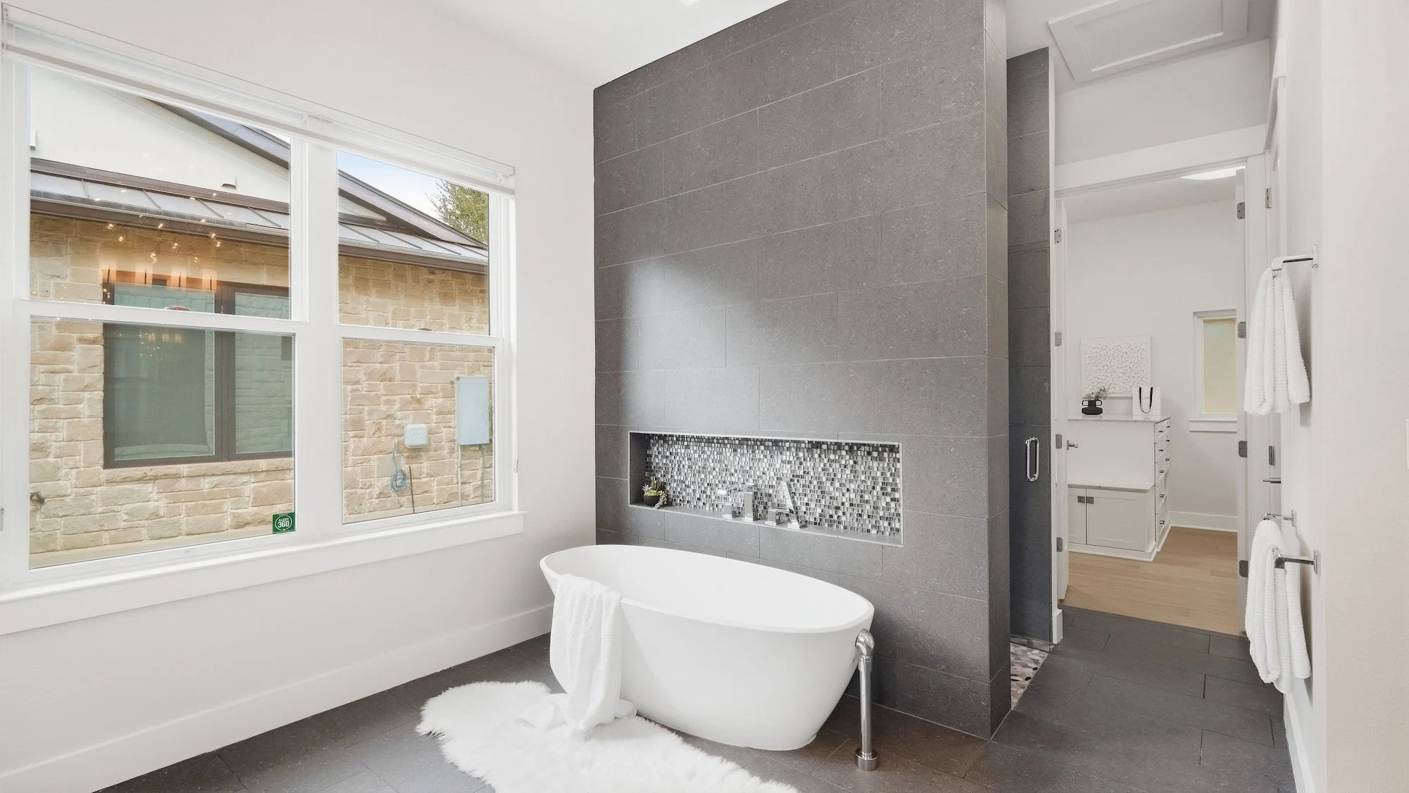 Primary bathroom staged with a freestanding tub and sheepskin rug before a gray tile wall with mosaic niche.