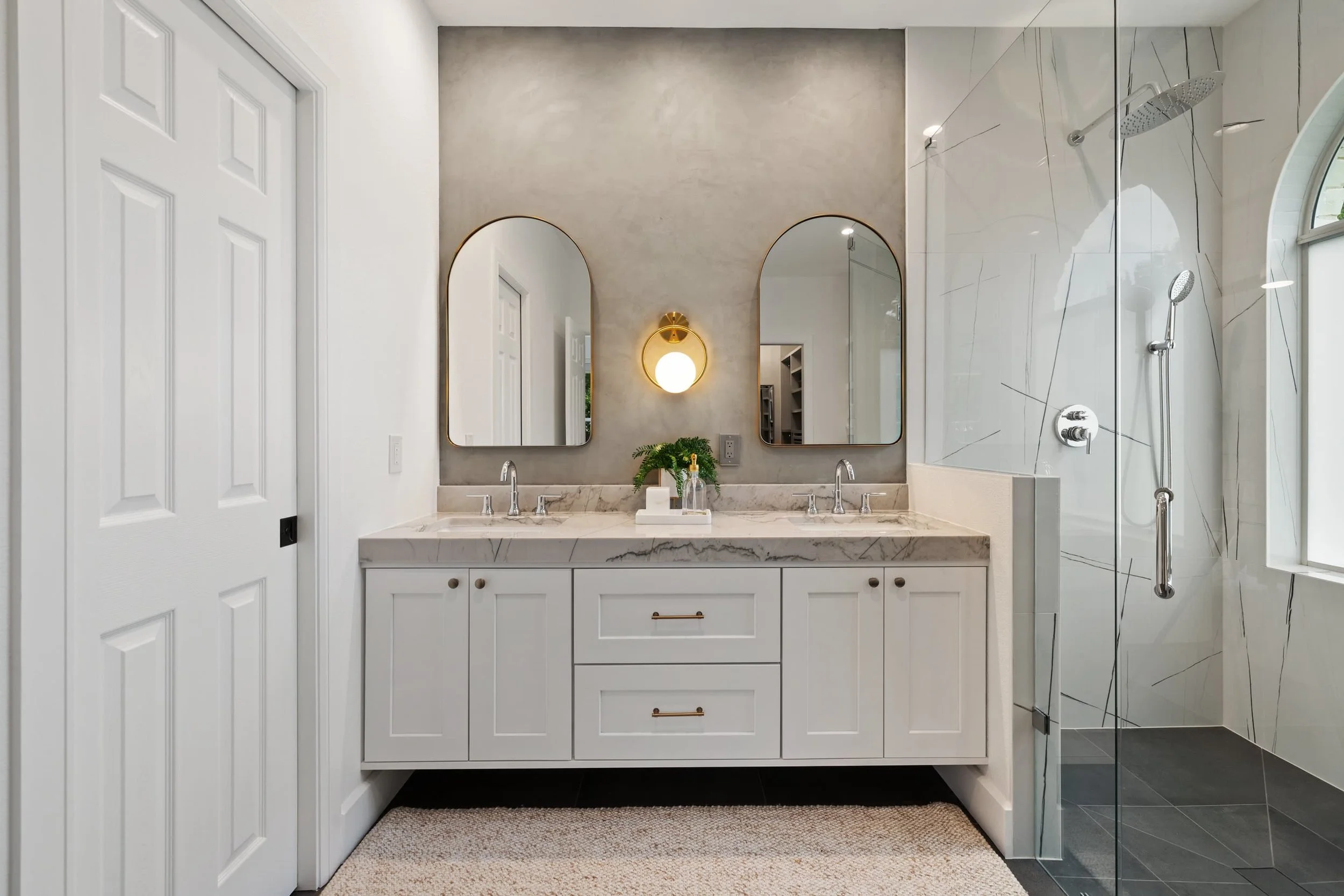 Modern bathroom with white double vanity, marble countertop, arched gold-framed mirrors, wall-mounted gold sconces, glass shower enclosure, and dark tile floor.