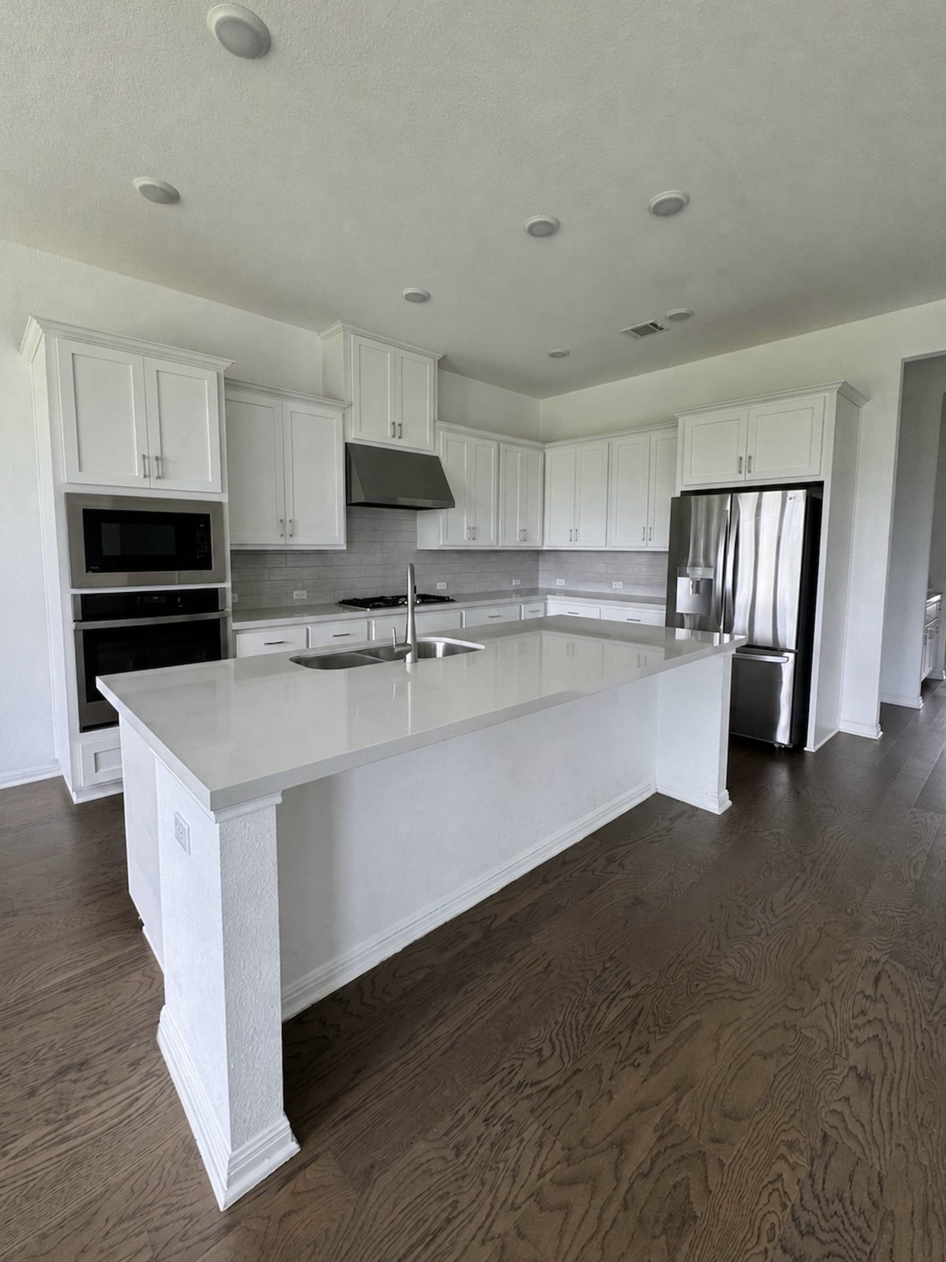 Vacant kitchen with white shaker cabinetry, a gray subway tile backsplash, stainless steel appliances including a built-in microwave and double oven, and a large quartz-topped center island with a sink.
