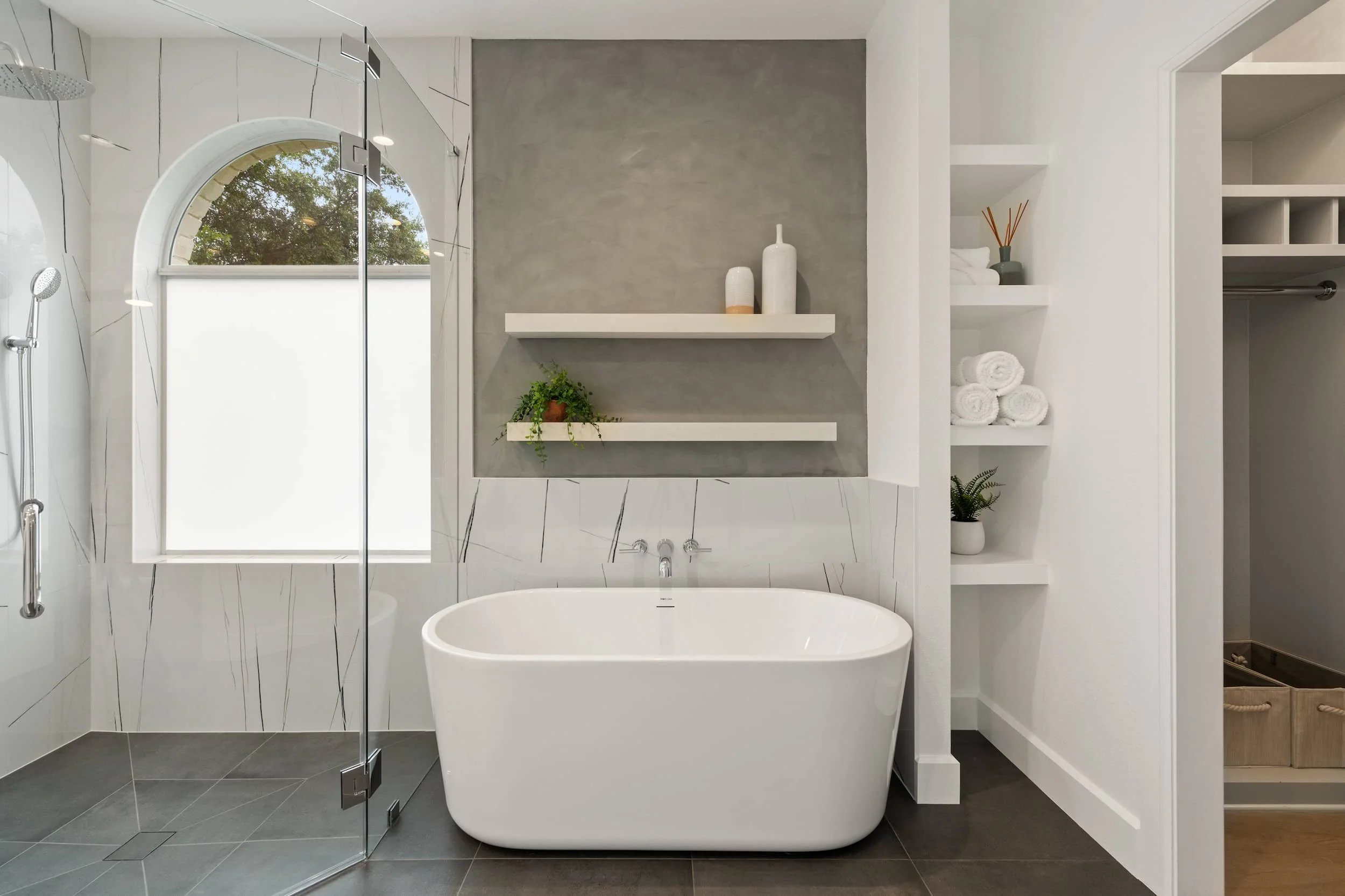 Bathroom with freestanding white oval bathtub, textured gray accent wall, floating white shelves, arched window, and frameless glass shower enclosure.