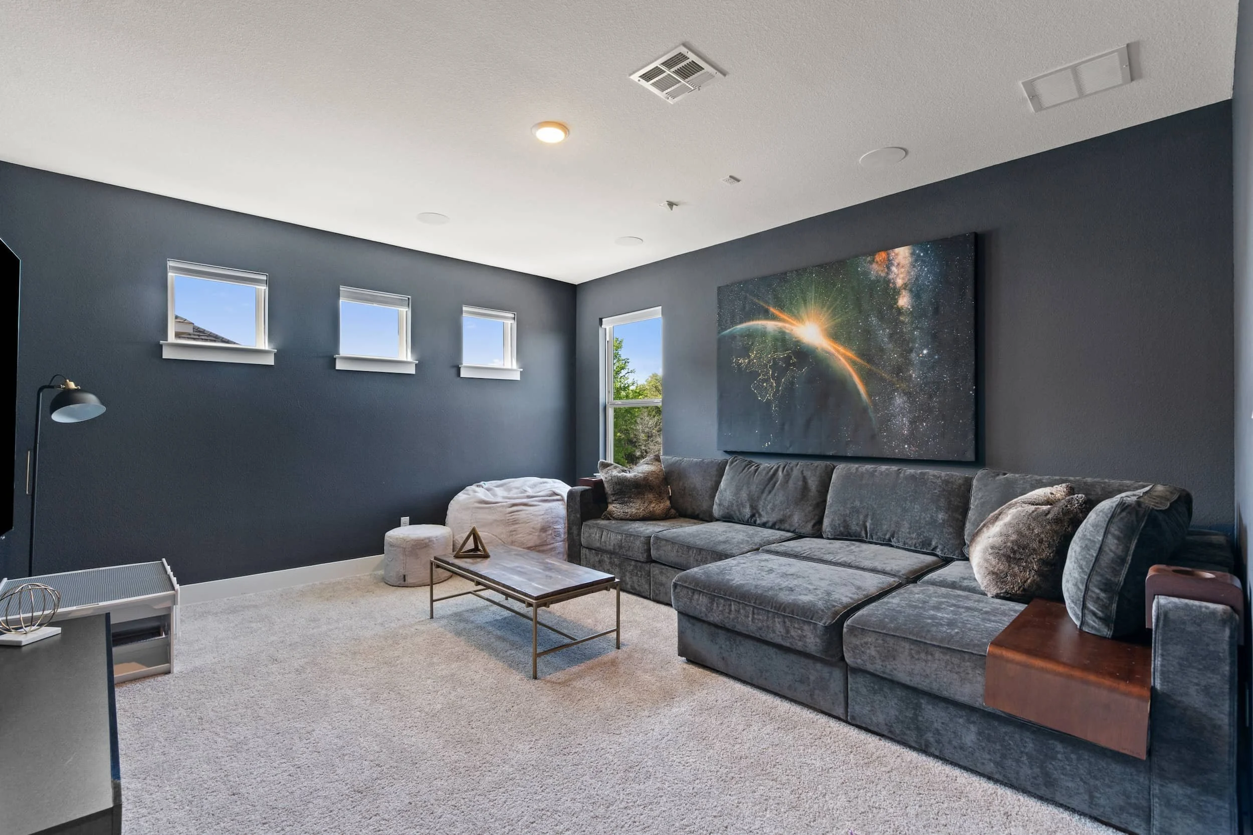 Staged media room with sectional sofa, dark accent walls, modern art, soft carpet, cozy seating layout.