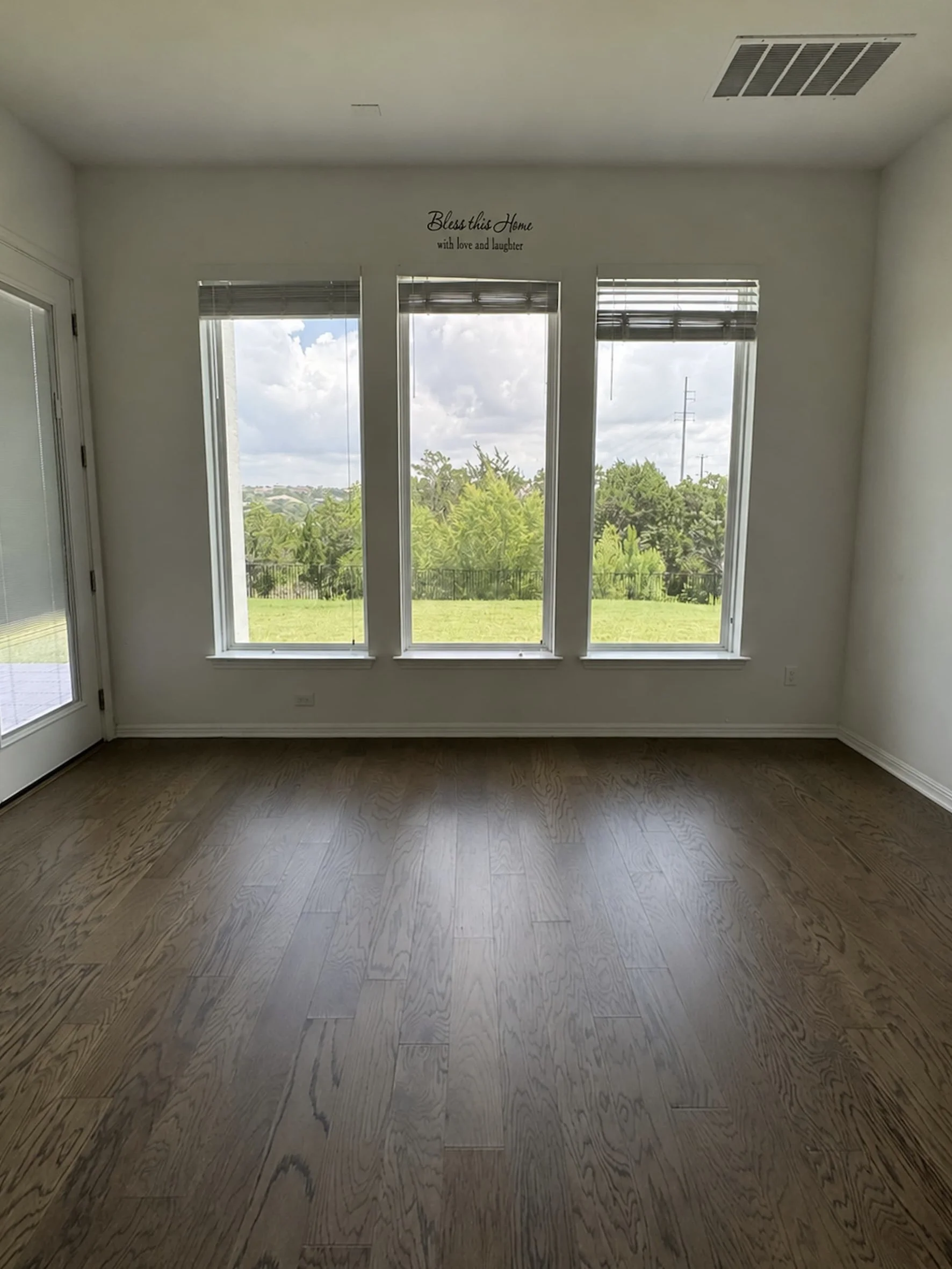 Empty breakfast nook with three tall windows overlooking a grassy backyard, dark hardwood floors, a glass door to the patio, and a decorative wall decal above the window trim.