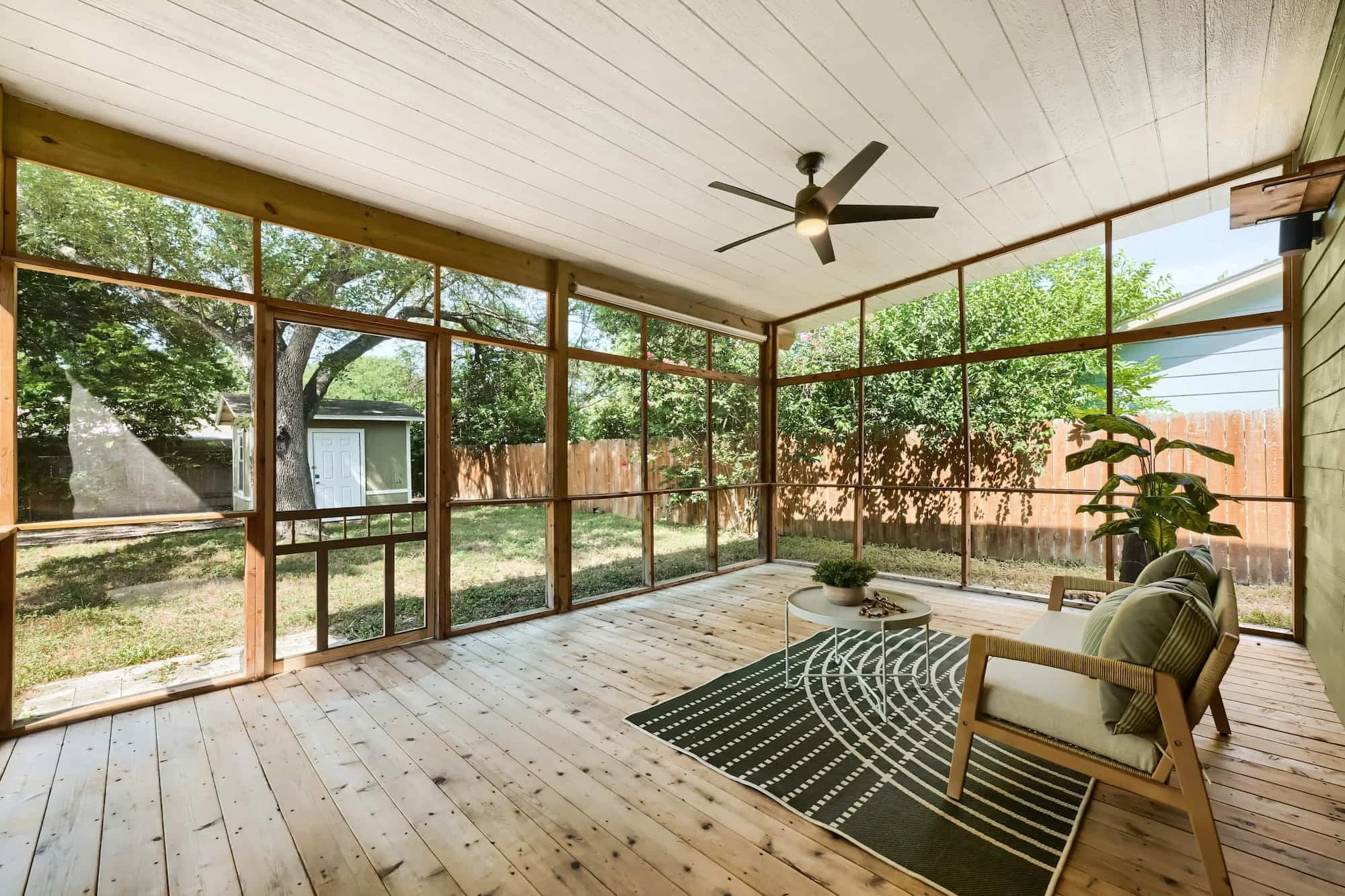 Staged screened-in porch with a wood-framed armchair and patterned rug on a plank deck, overlooking a fenced backyard.