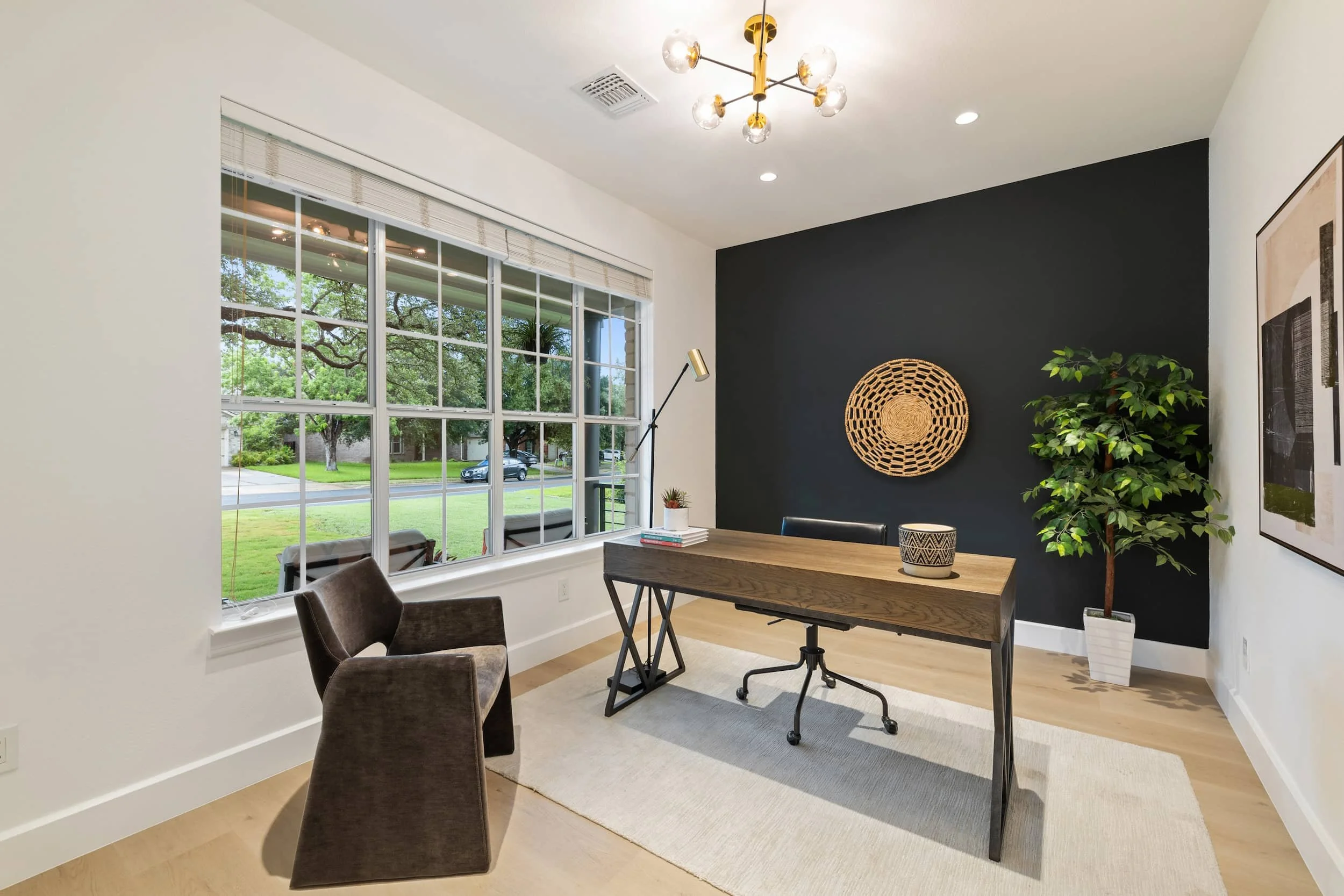 Home office with black accent wall, woven circular art, dark wood desk, leather chair, large windows overlooking trees and street, and potted plant in the corner.