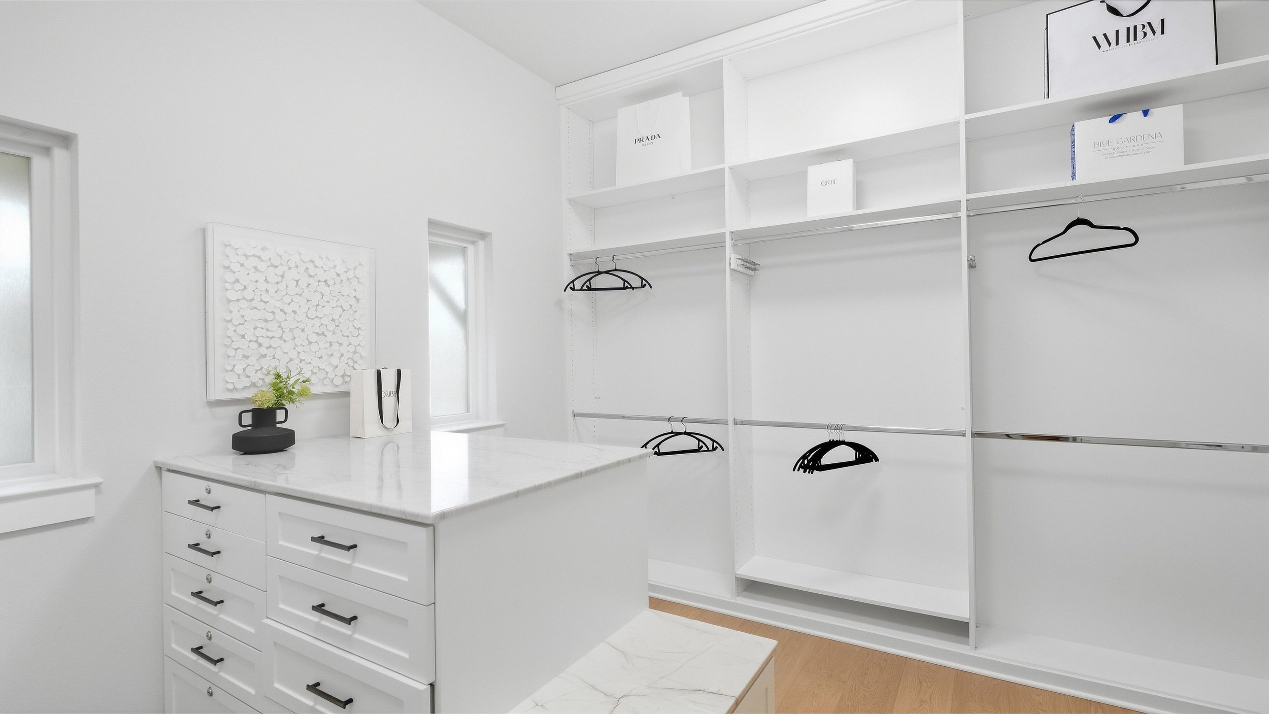 Primary closet staged with a marble-top dresser island, boutique shopping bags, and white textured wall art.