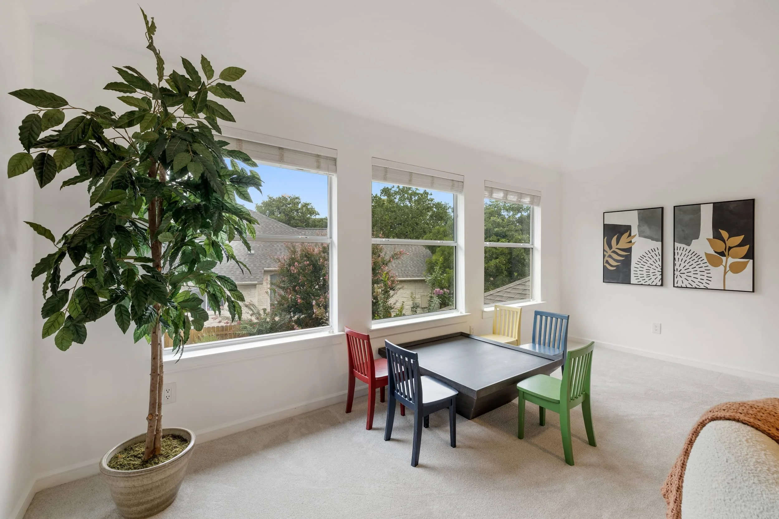 Modern kids' dining area with colorful chairs, dark table, large windows with tree views, faux ficus tree, and botanical wall art.