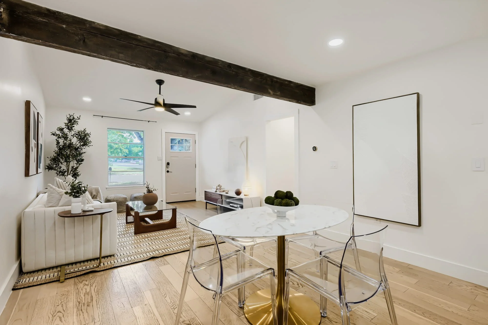 Staged dining area with a round marble table and clear acrylic chairs, open to the living room under an exposed wood beam.