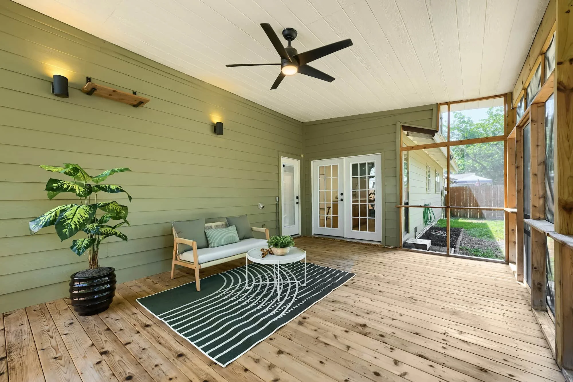 Staged screened-in porch with a cushioned loveseat and patterned rug on a wood deck, beside sage siding and french doors into the home.