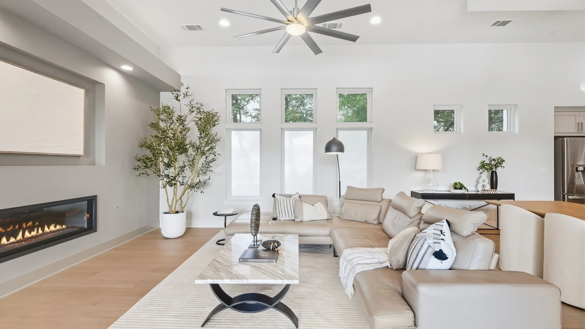 Living room staging with a cream sectional, potted olive tree, and tall windows beside a linear fireplace.