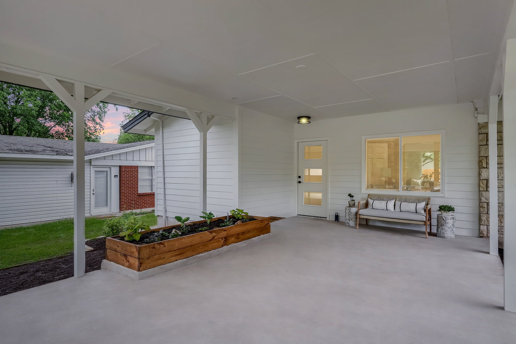 Covered rear patio with white siding, concrete floor, bench seating, potted plants, and a raised wooden garden bed.