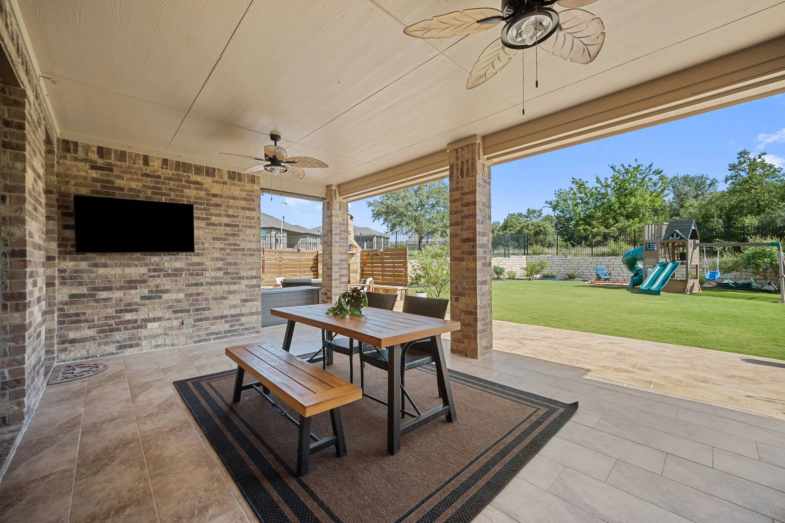 Covered patio staging with brick columns, wood dining set, tile flooring, ceiling fans, outdoor TV, spacious backyard and lawn view.