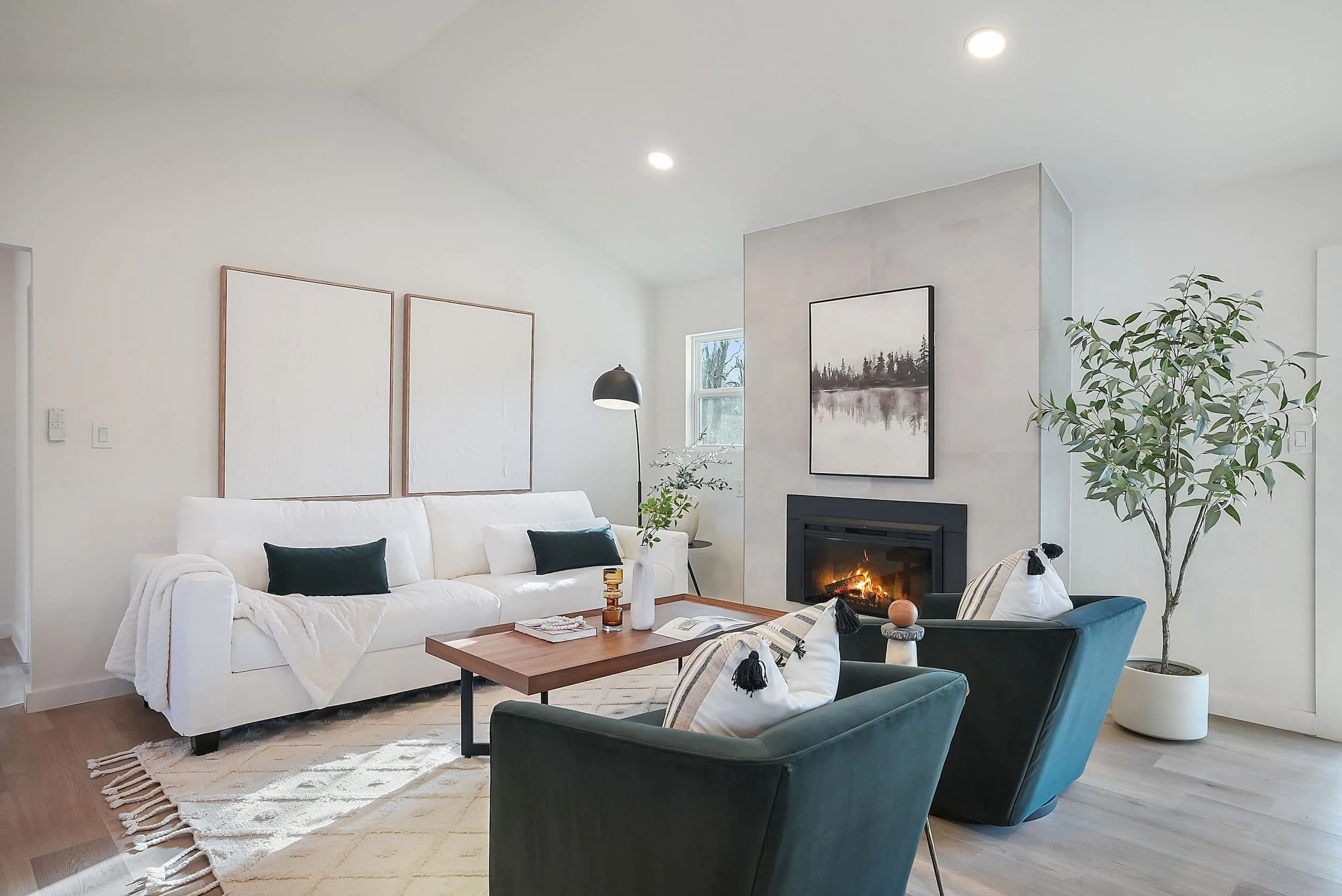Cozy neutral family room: white couch, green velvet chairs, walnut table with decor, tall textured fireplace with burning logs and forest print, arc floor lamp, and high ceiling.