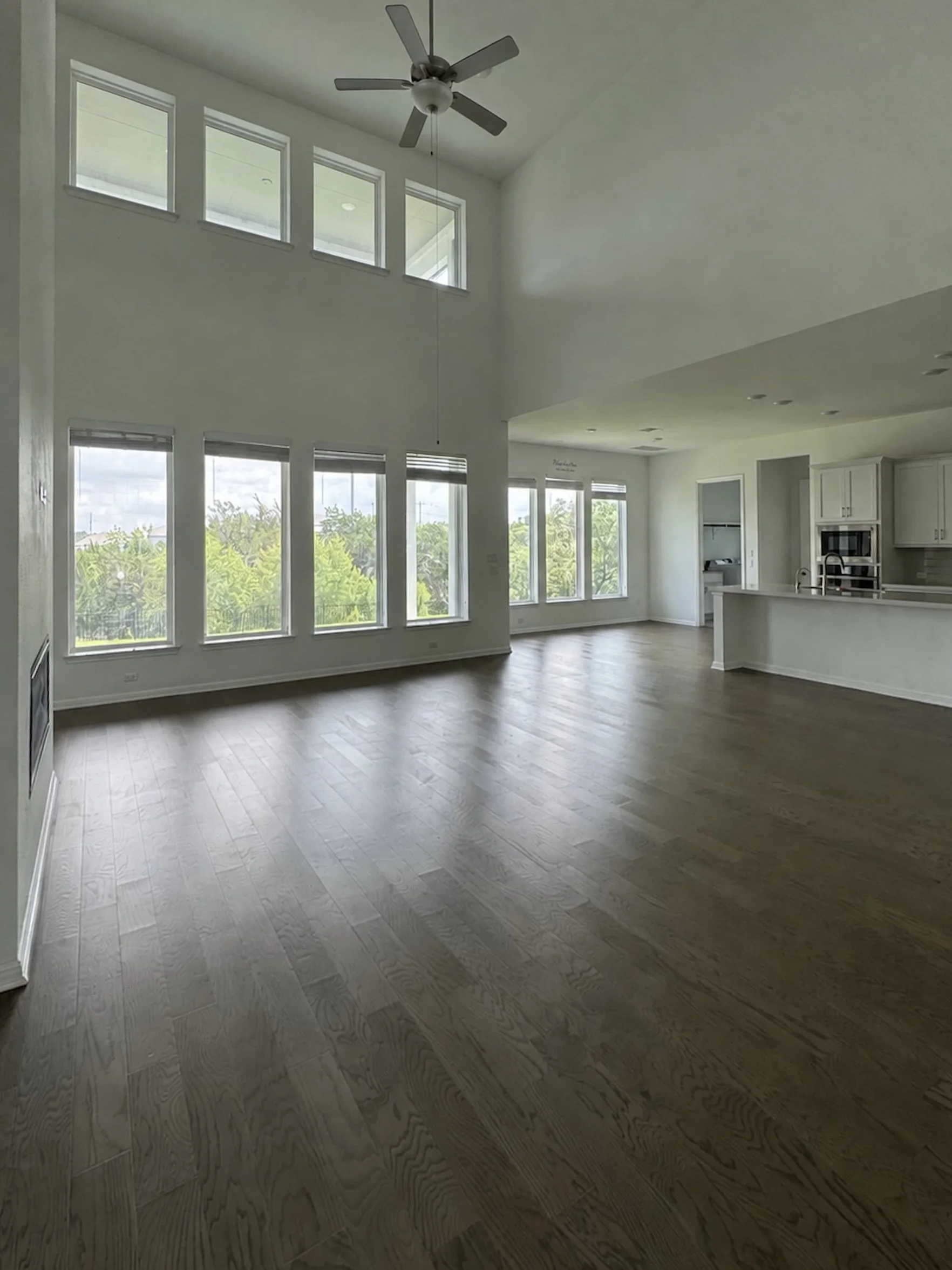 Vacant two-story great room with a wall of tall windows, dark hardwood flooring, and a ceiling fan, opening directly into the kitchen with a view of the white cabinetry and island beyond.