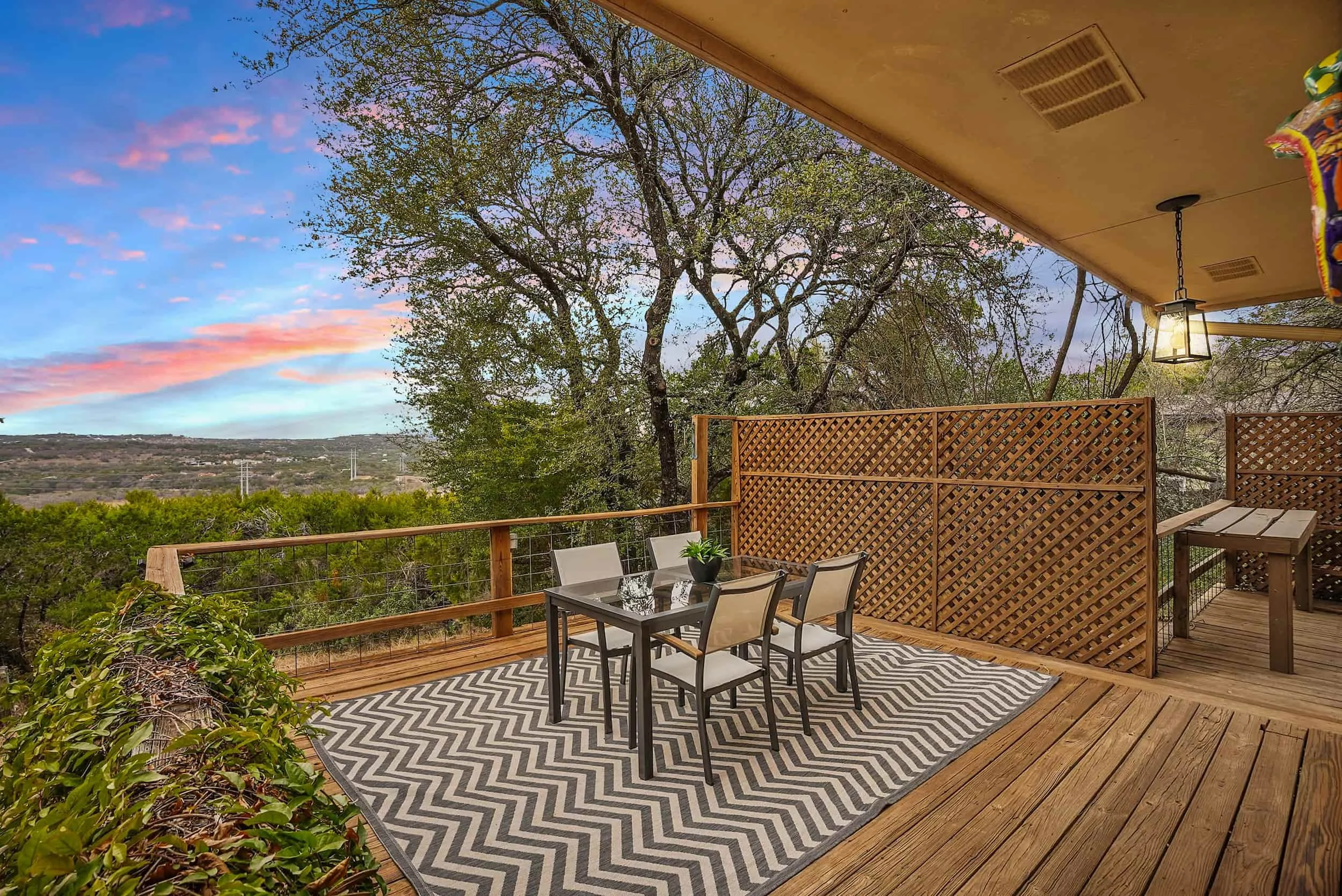 Spacious covered deck with outdoor dining set, privacy screen, and sweeping views of trees and hills under a colorful sunset sky.