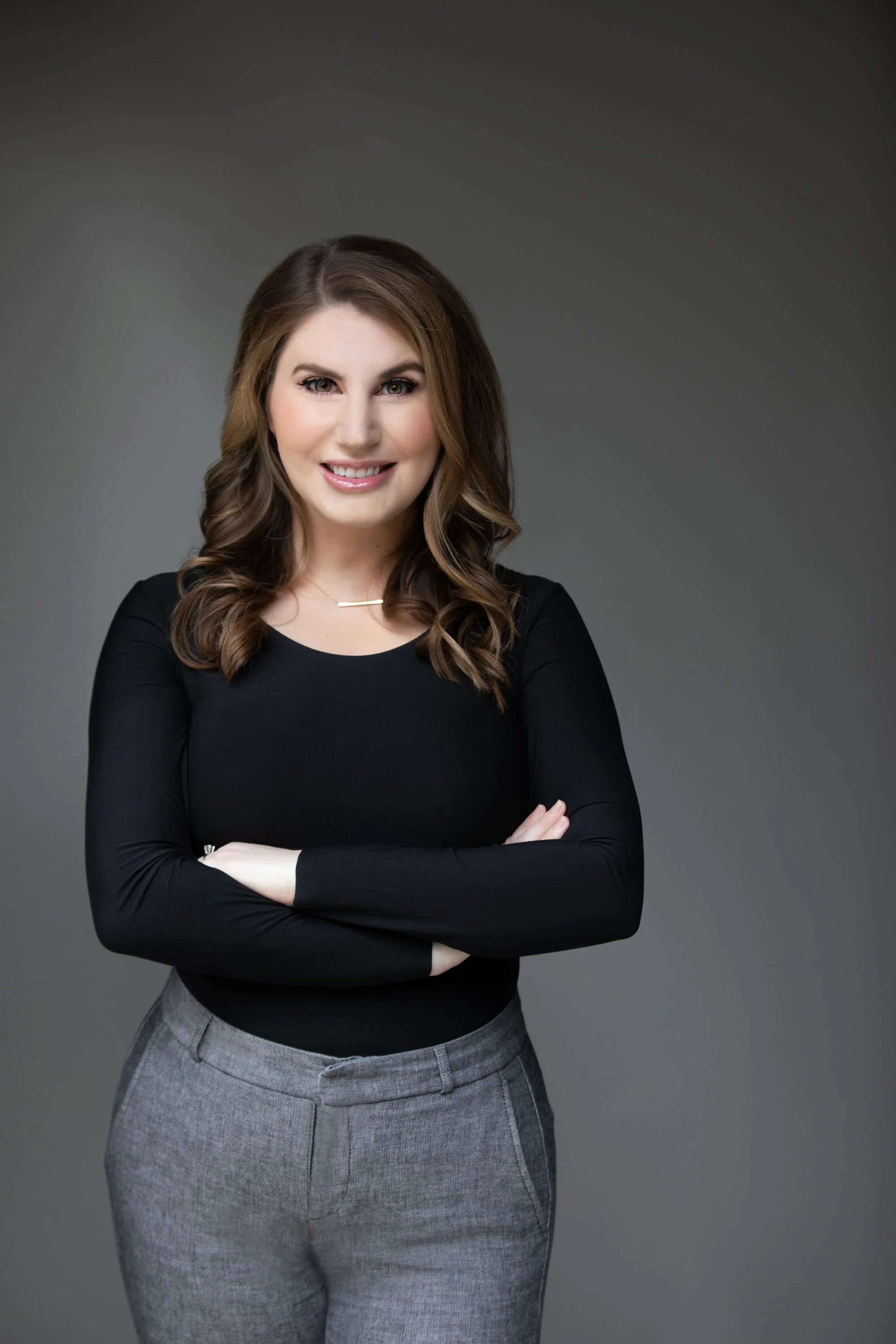 A woman with wavy brown hair, smiling, wearing a black long sleeve shirt and gray dress pants, standing against a dark gradient background.