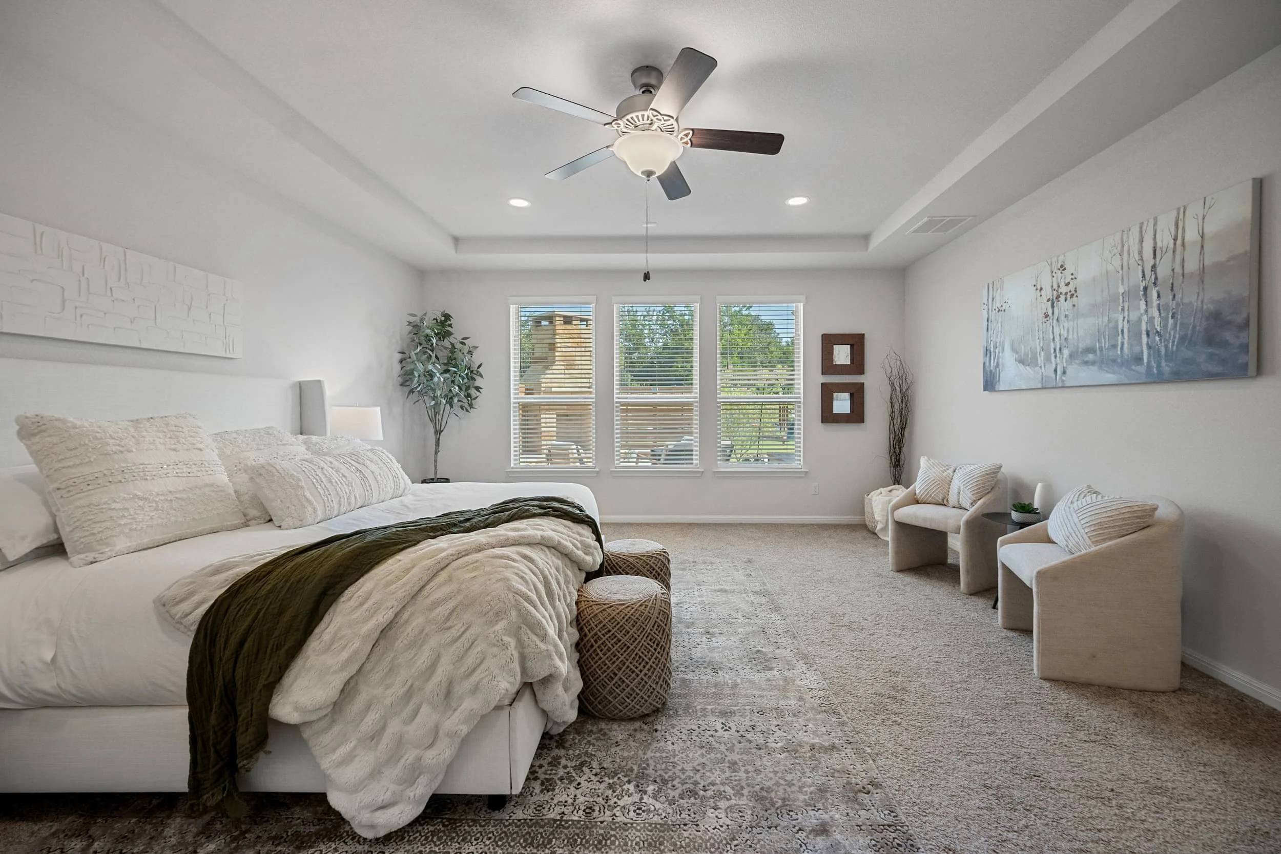 Bright master suite with neutral carpet, olive green throw, woven ottomans, and a tray ceiling with a dark wood fan.