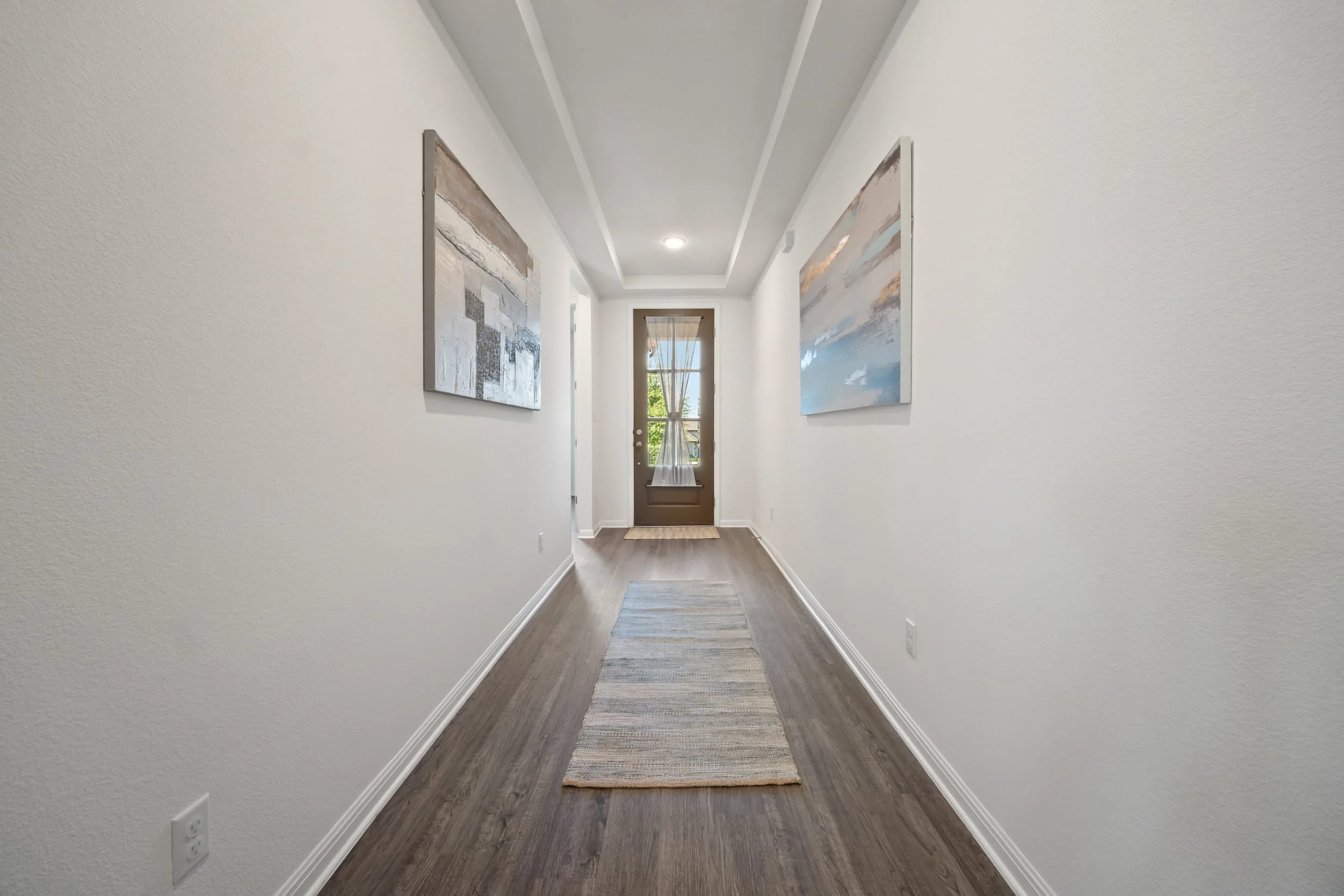 Bright modern hallway with white walls, wood-look floors, abstract art, and runner rug leading to glass door in staged home.