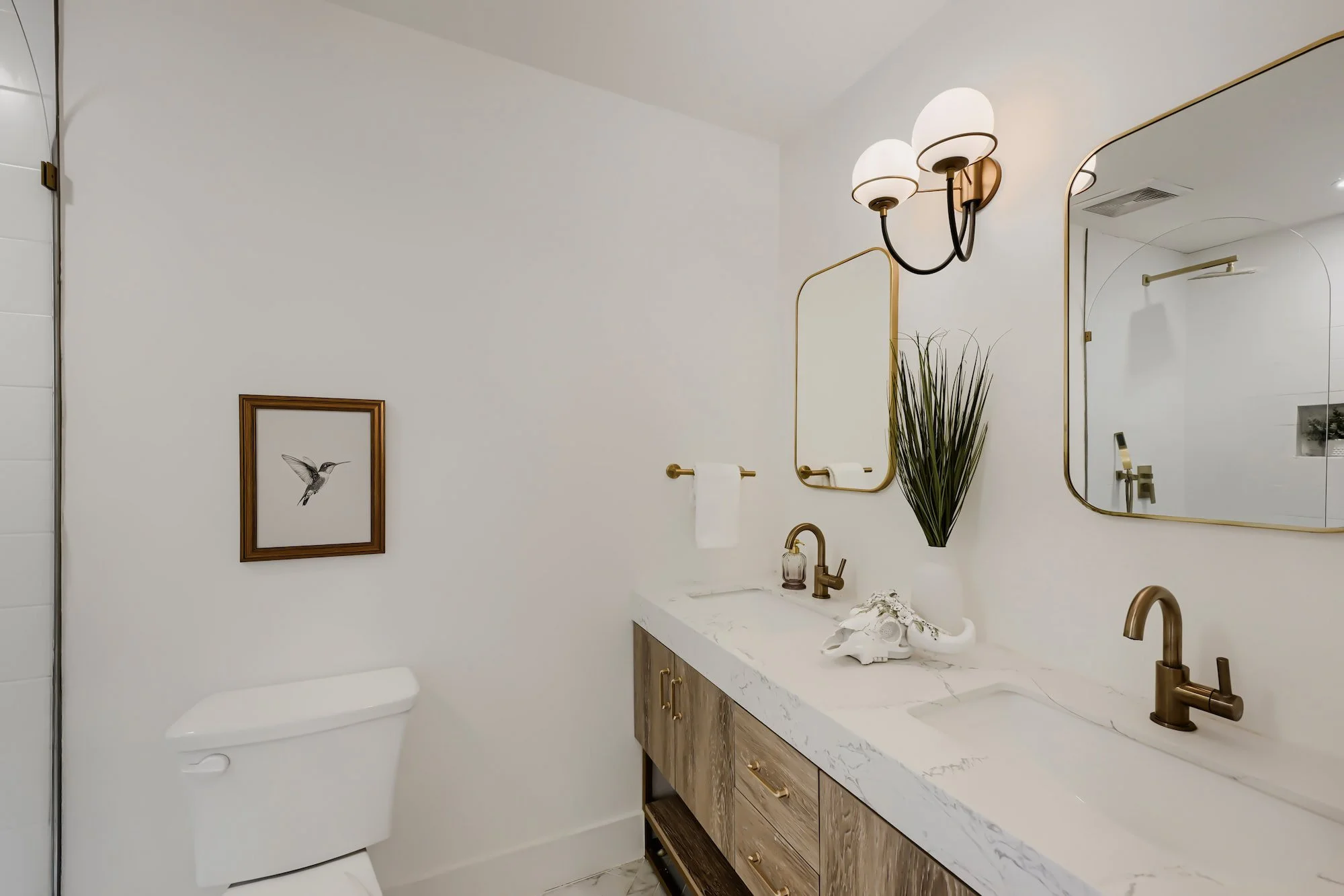 Staged primary bathroom with a wood double vanity, white marble counter, brass faucets, and arched mirrors under a globe sconce.