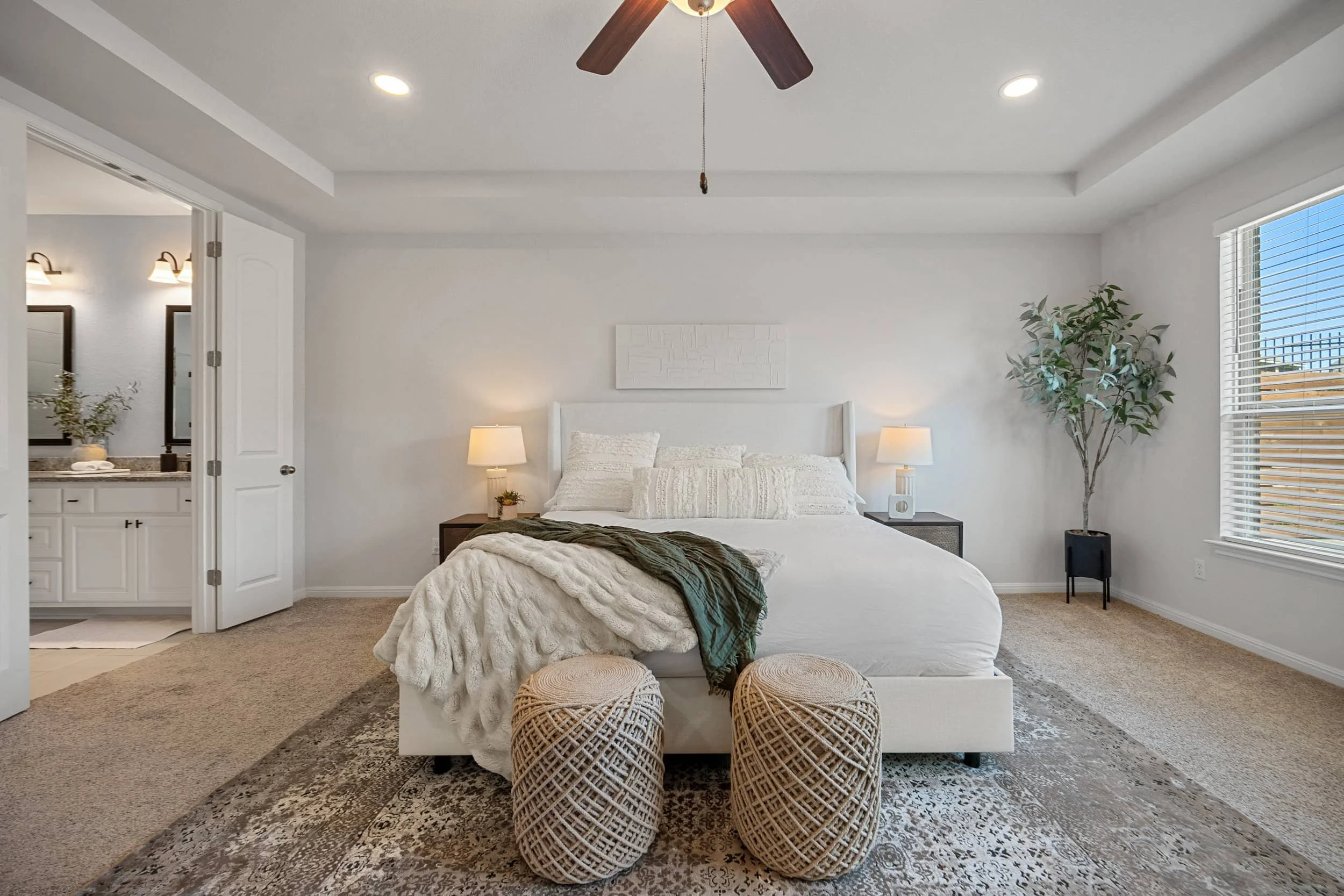 Staged primary bedroom with white bedding, woven ottomans, a tray ceiling, and an attached bathroom in Austin.