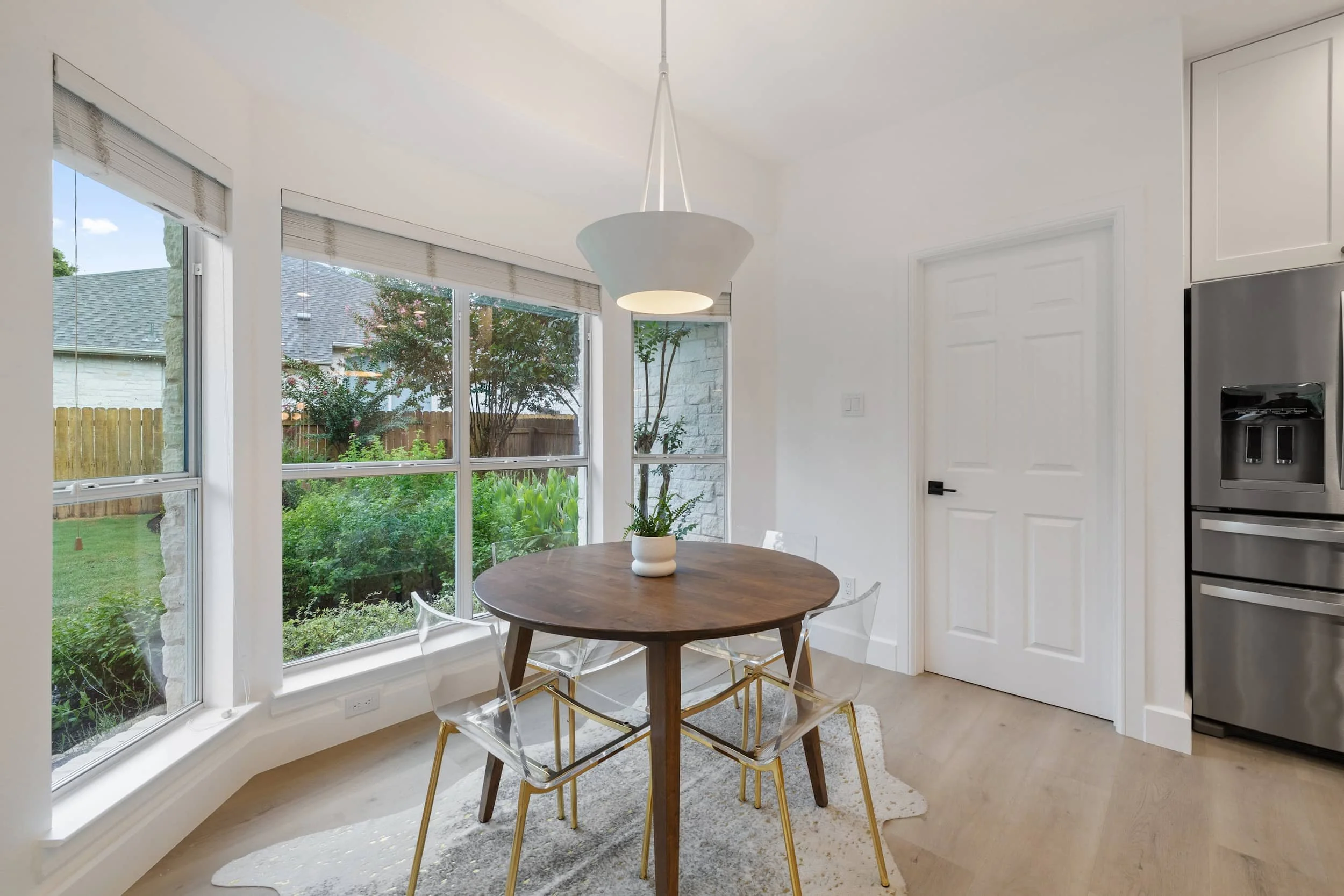 Light-filled breakfast nook with a round wooden table, clear acrylic chairs with gold legs, pendant light, and large windows overlooking a landscaped backyard.