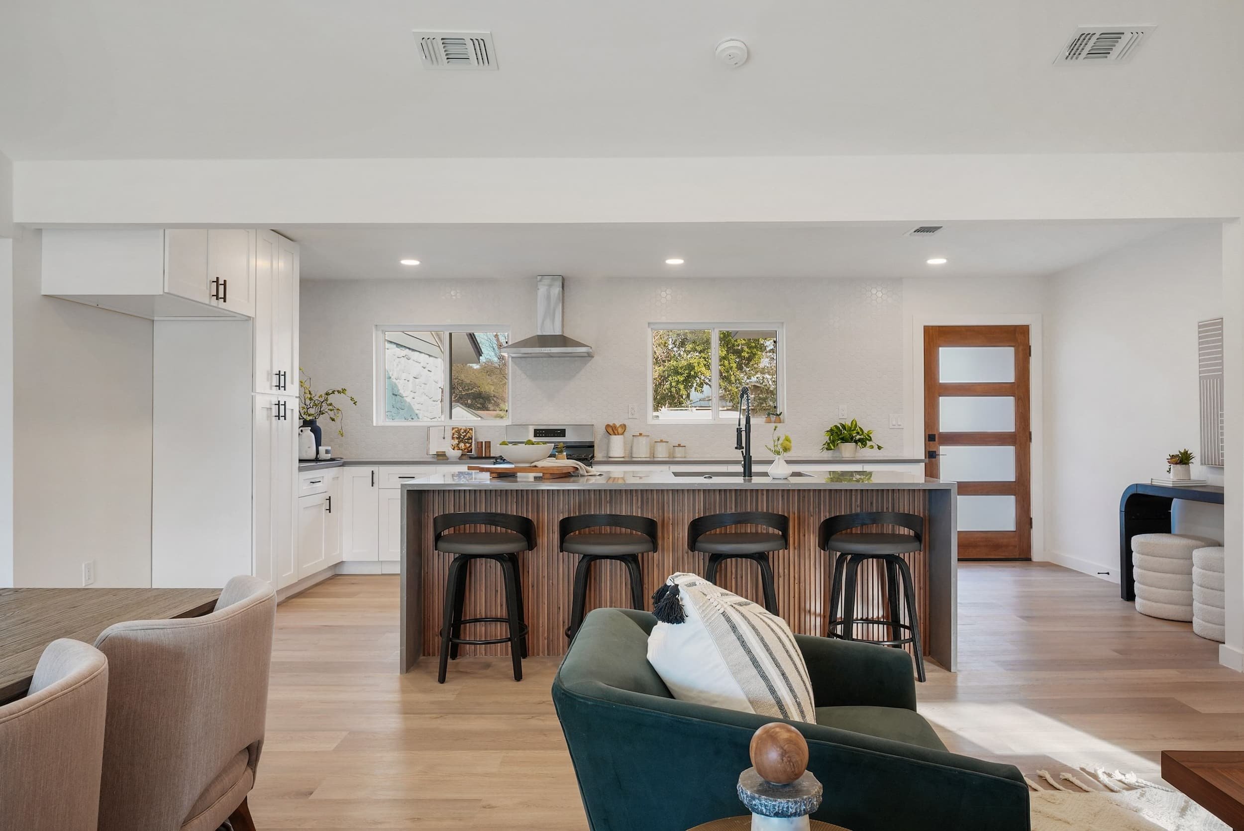 Modern open-concept kitchen with a wood-slat island, white cabinetry, and a green velvet armchair in the foreground.
