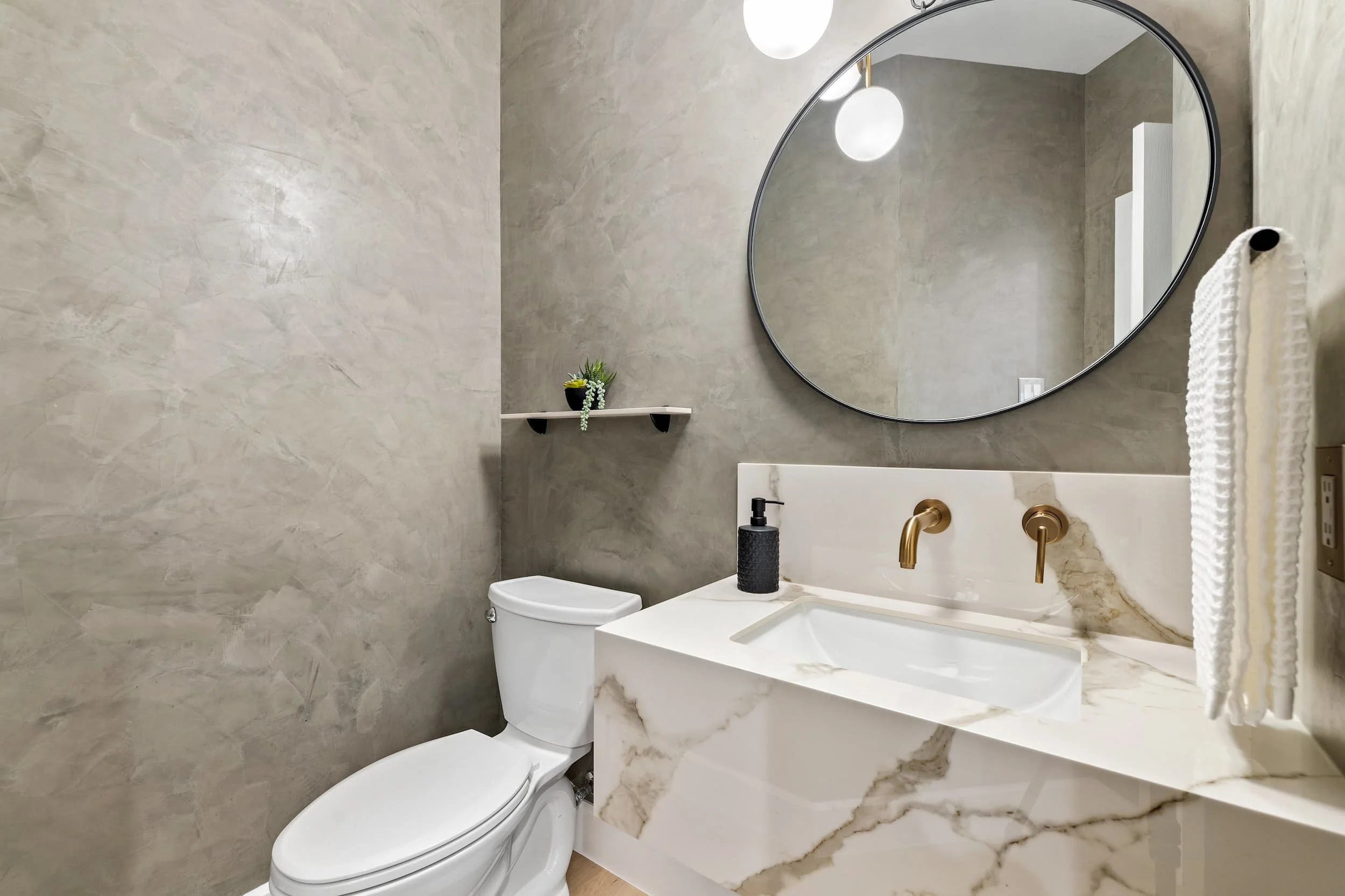 Stylish powder room with a floating marble vanity, gold wall-mounted faucet, round black-framed mirror, globe lighting, textured gray walls, and a compact toilet.