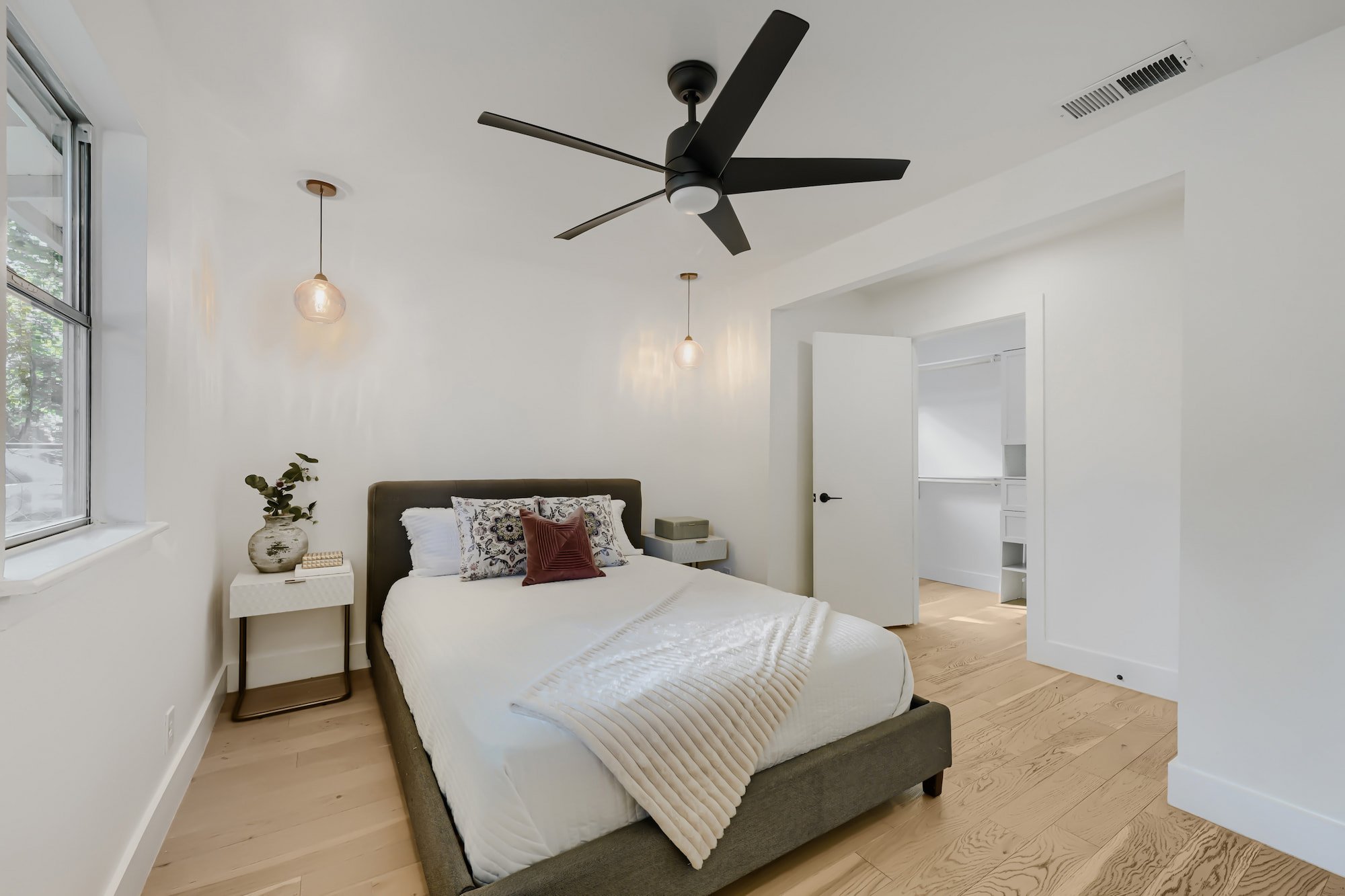 Staged primary bedroom with an upholstered bed, brass pendants, and a nightstand, beside an open walk-in closet.