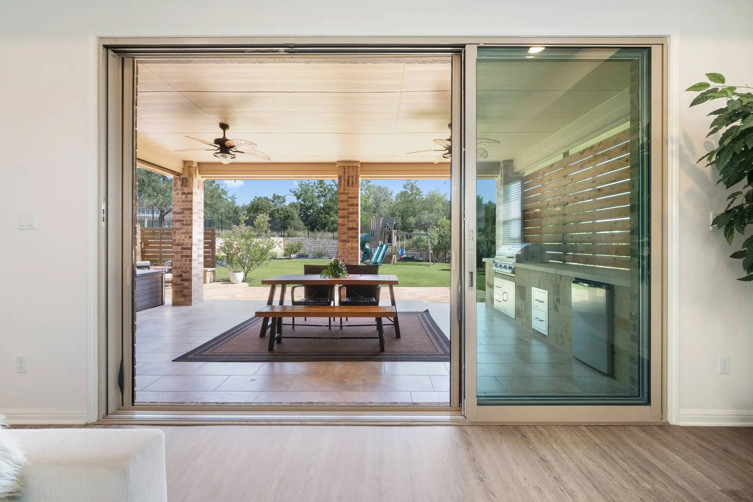 Indoor-outdoor living with sliding glass doors, covered brick patio, wood dining set, tile flooring, built-in grill, open backyard view in Austin