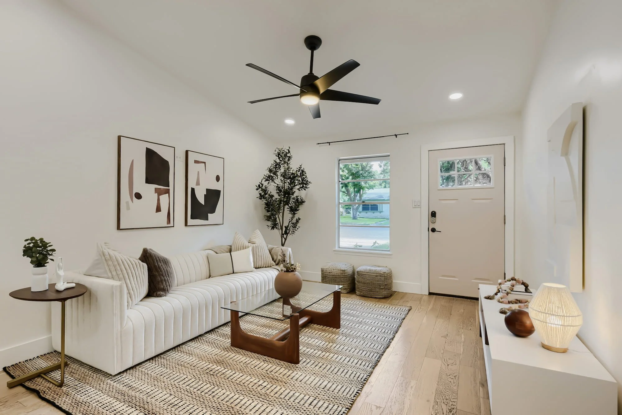Staged living room with a channel-tufted white sofa, woven poufs, and abstract art, facing a light-filled front entry.