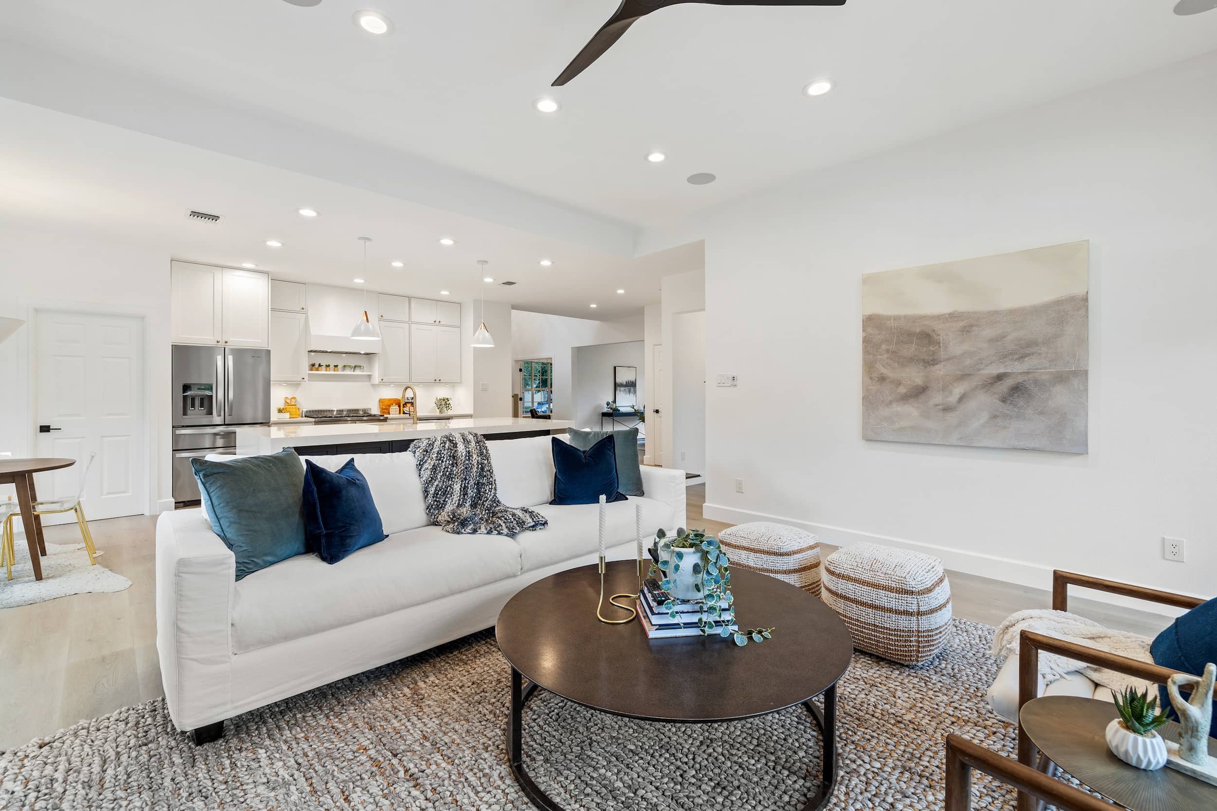Open-concept living room with white sofa and coffee table, flowing into a modern kitchen with white cabinets, stainless steel appliances, and pendant lighting.