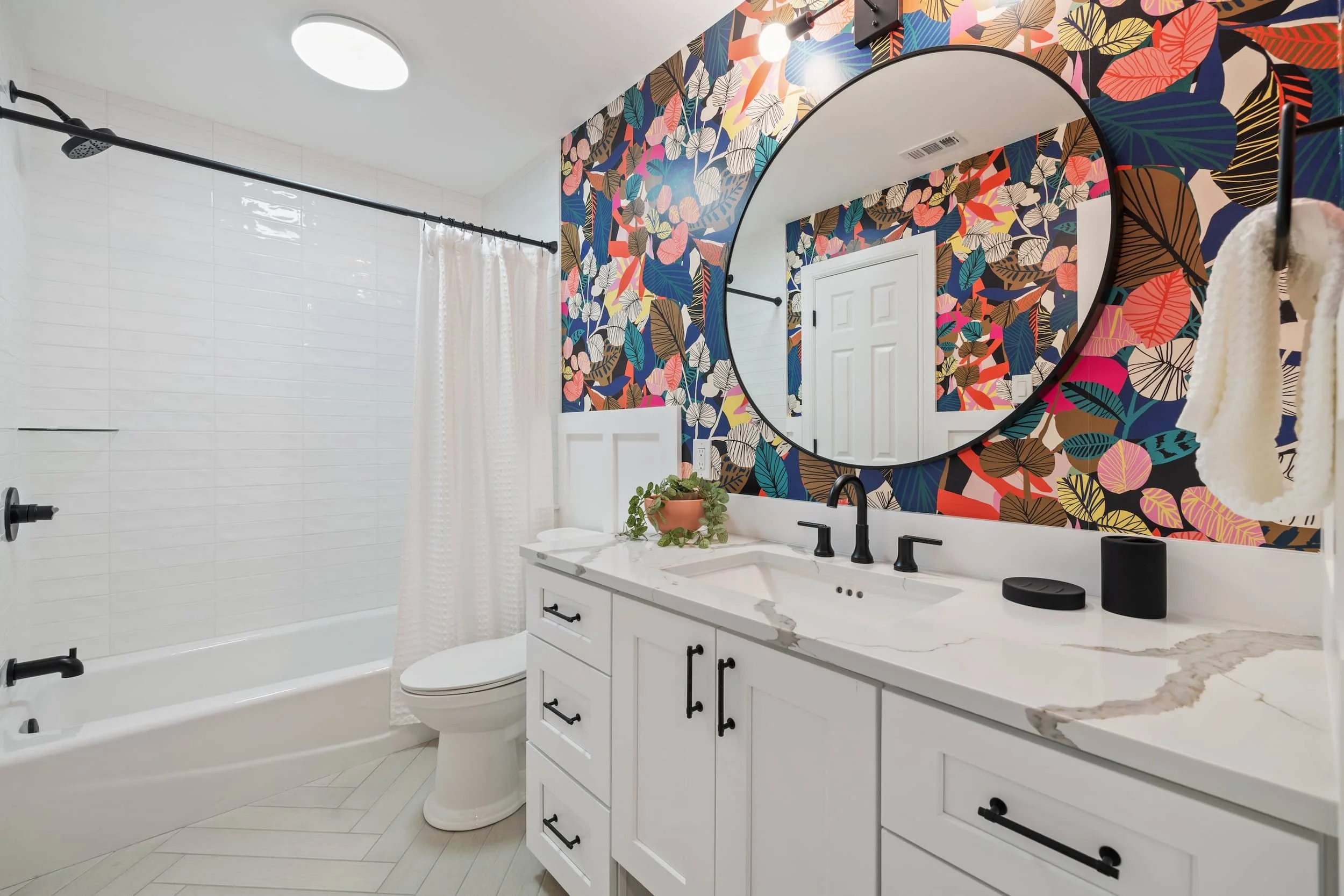 Maximalist bathroom with a white vanity, black fixtures, round mirror, and bold floral wallpaper accent wall, alongside a white tiled tub and shower.