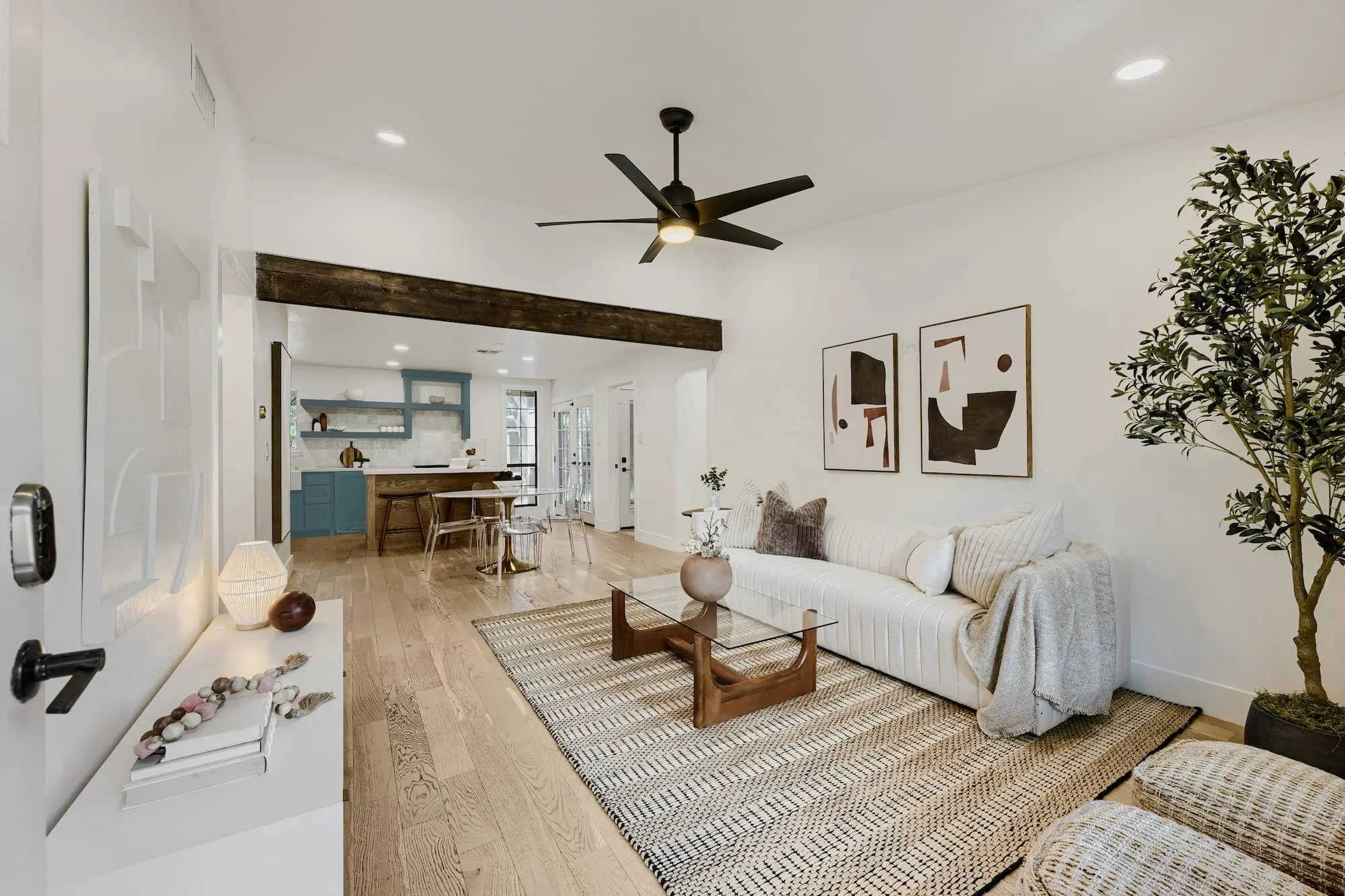 Staged living room with a tufted ivory sofa and wood-and-glass coffee table on a jute rug, opening to a teal kitchen beyond.