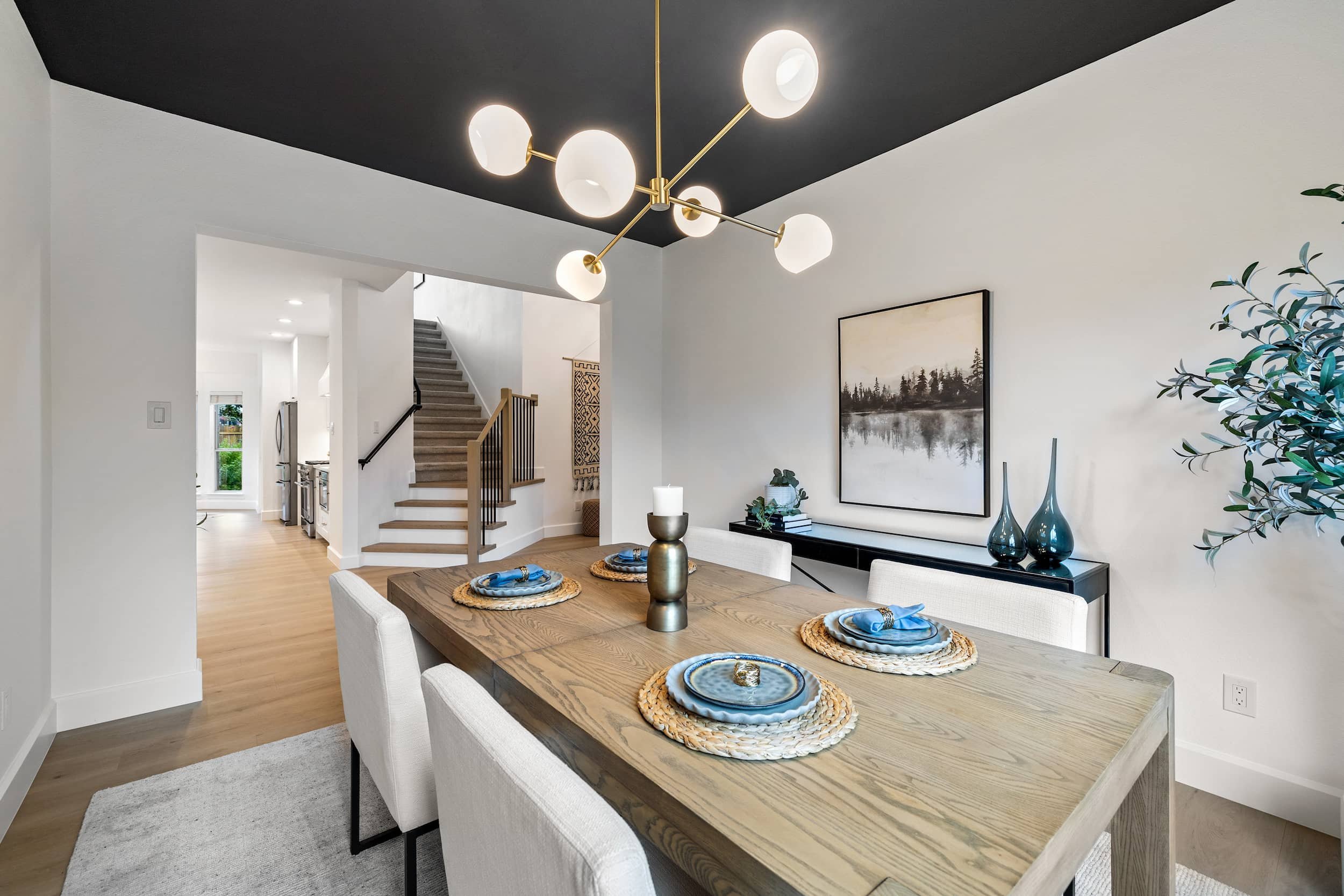 Dining room with dark ceiling, gold chandelier, long wooden table, beige upholstered chairs, black console table with vase and books.