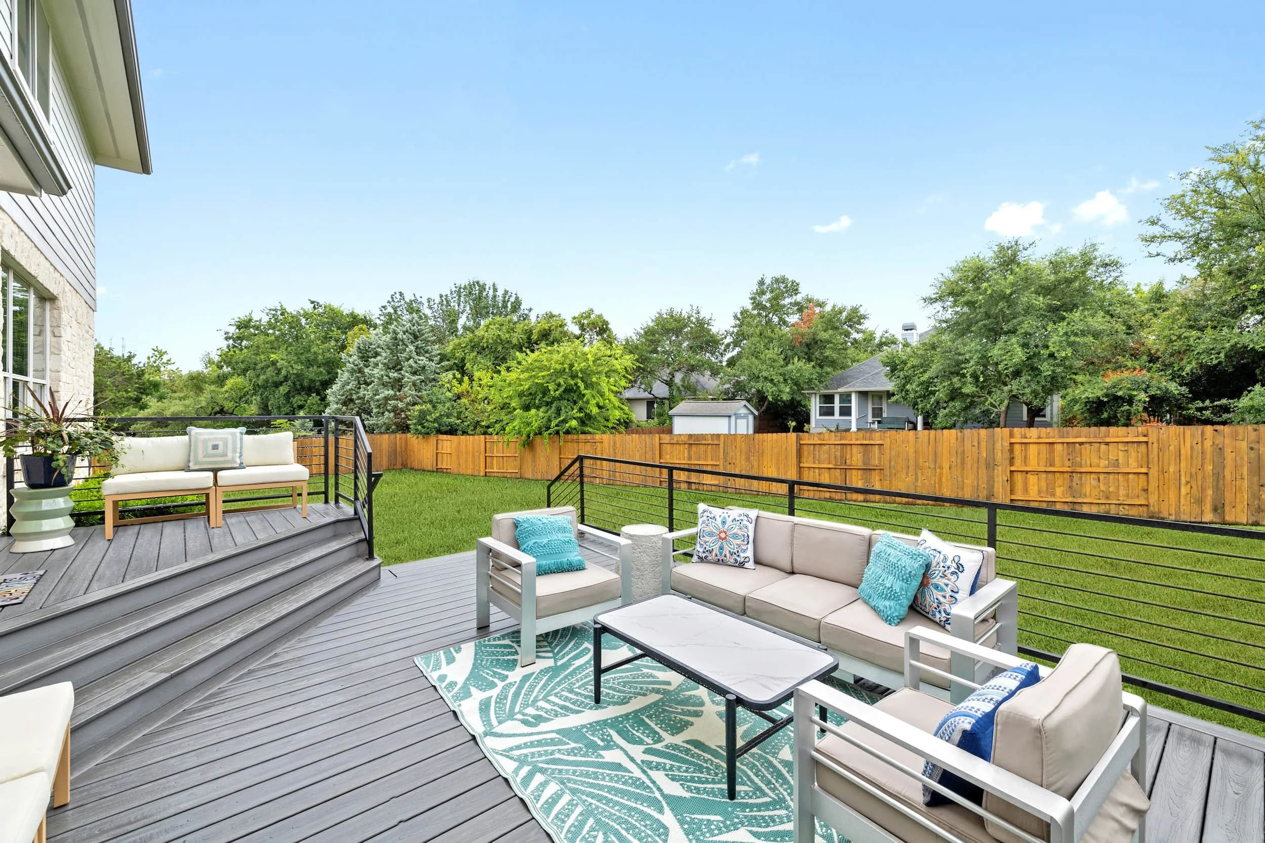 Spacious backyard deck with outdoor sofa and chairs, coffee table, and railing, overlooking a grassy fenced yard with trees.